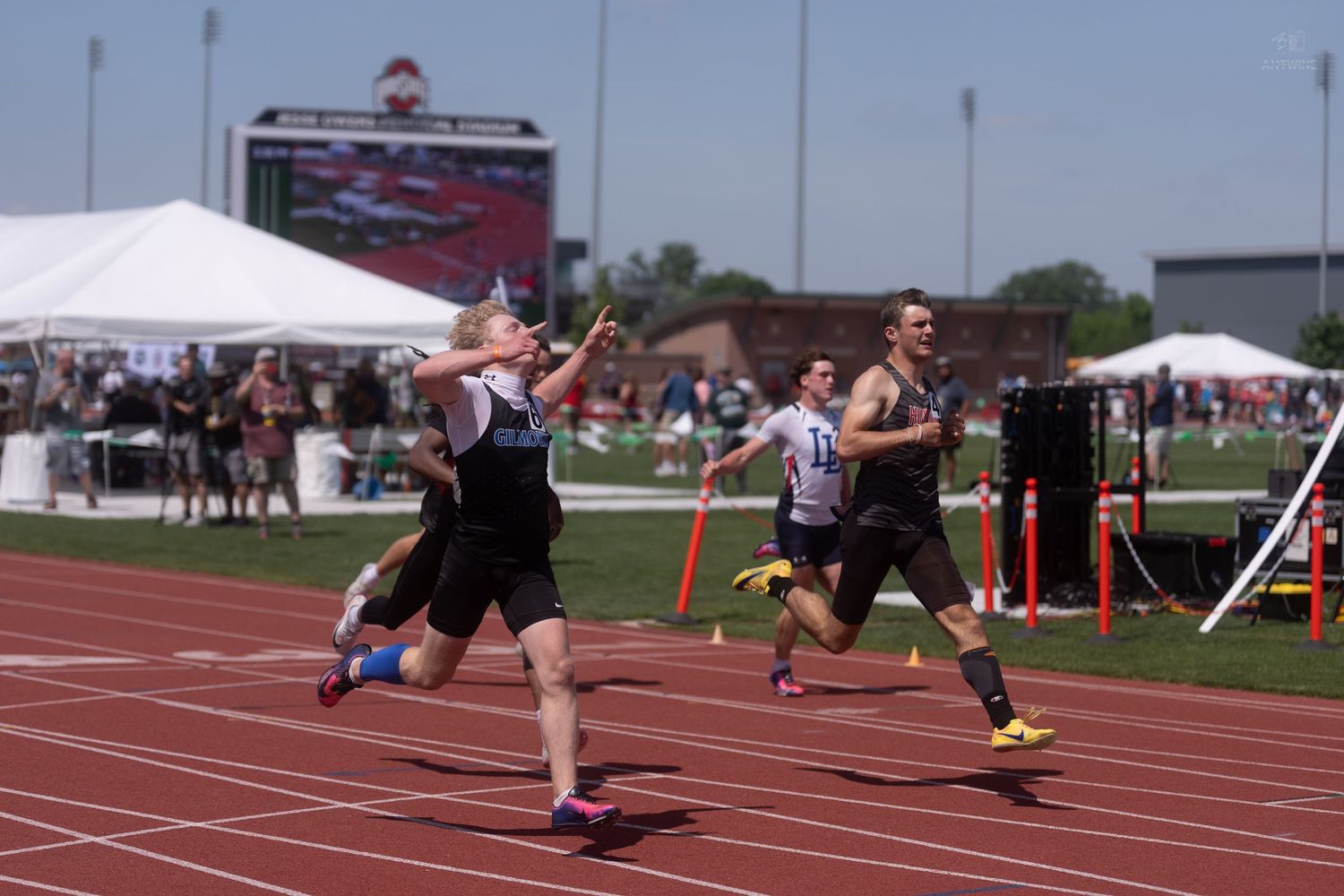 Runner crosses the finish line with arms raised at an outdoor track and field championship meet.