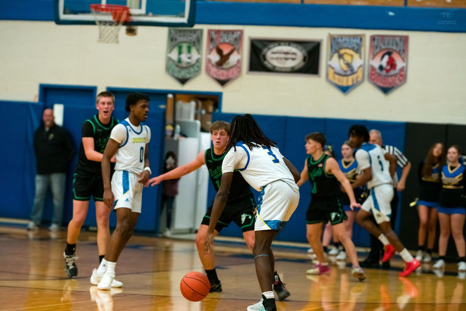 A blue-uniformed player dribbles past a green-jersey defender beneath championship banners in a high school gymnasium.