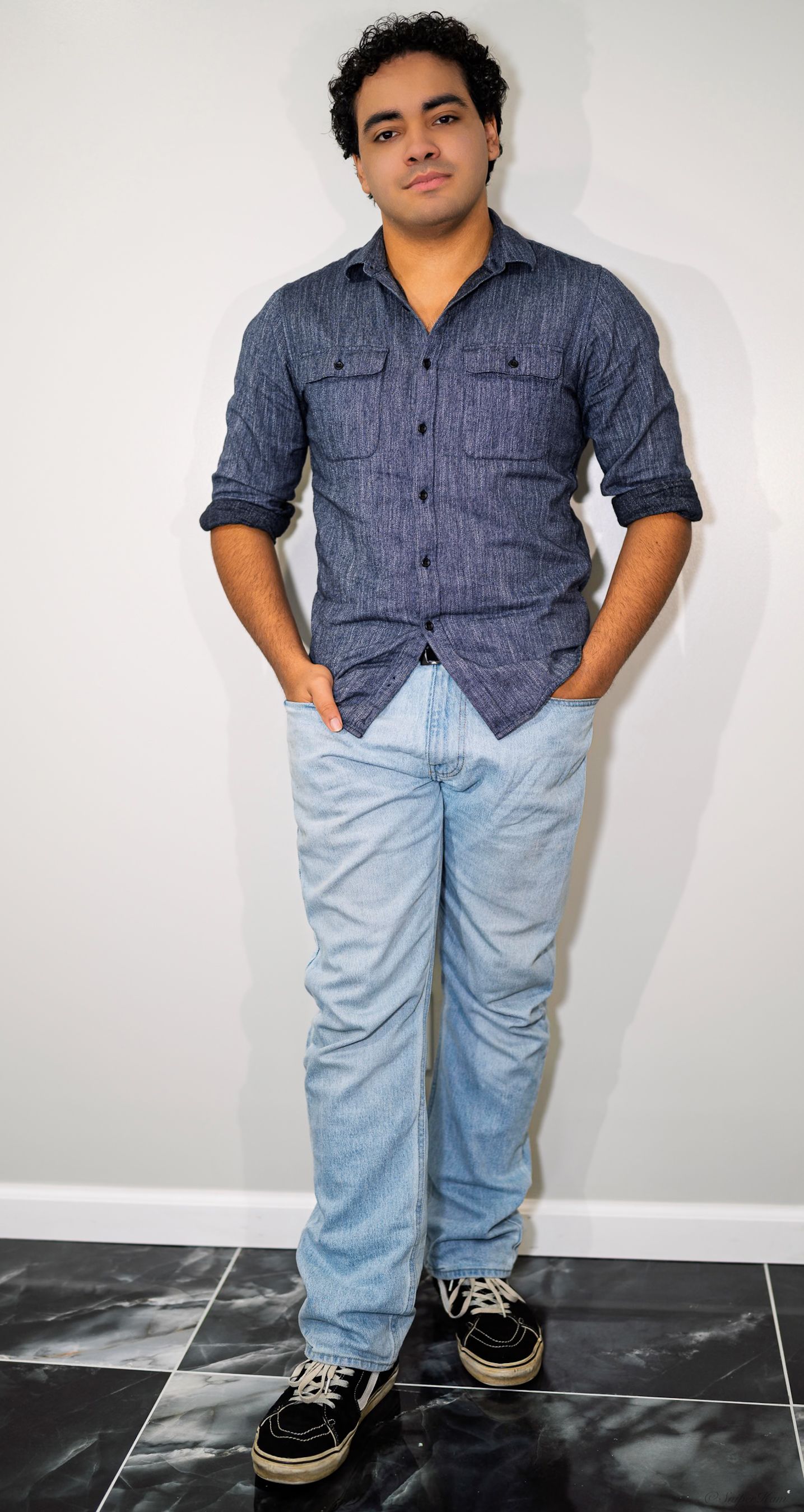 Young man in blue chambray shirt and light jeans standing against white wall.