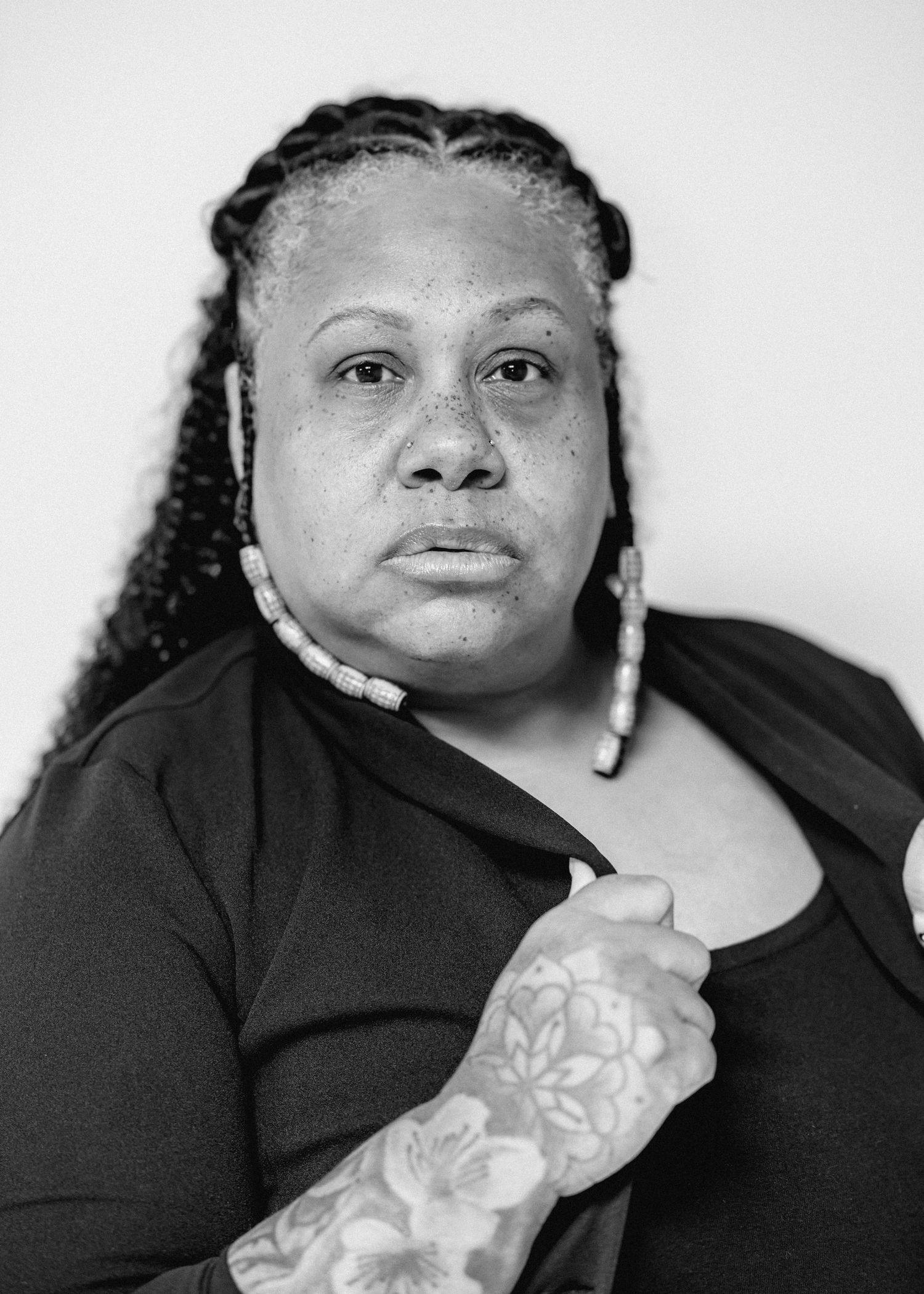Black woman with braided hair and beaded earrings showing floral arm tattoo, monochrome portrait photo.