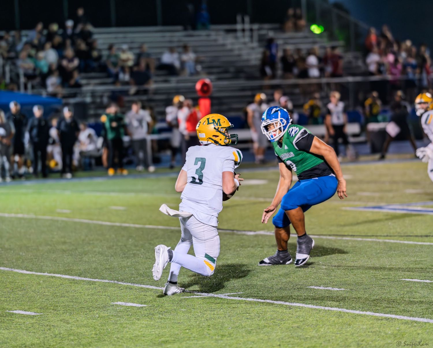 Football player in white uniform runs as a defender in green and blue uniform moves in to make a tackle.