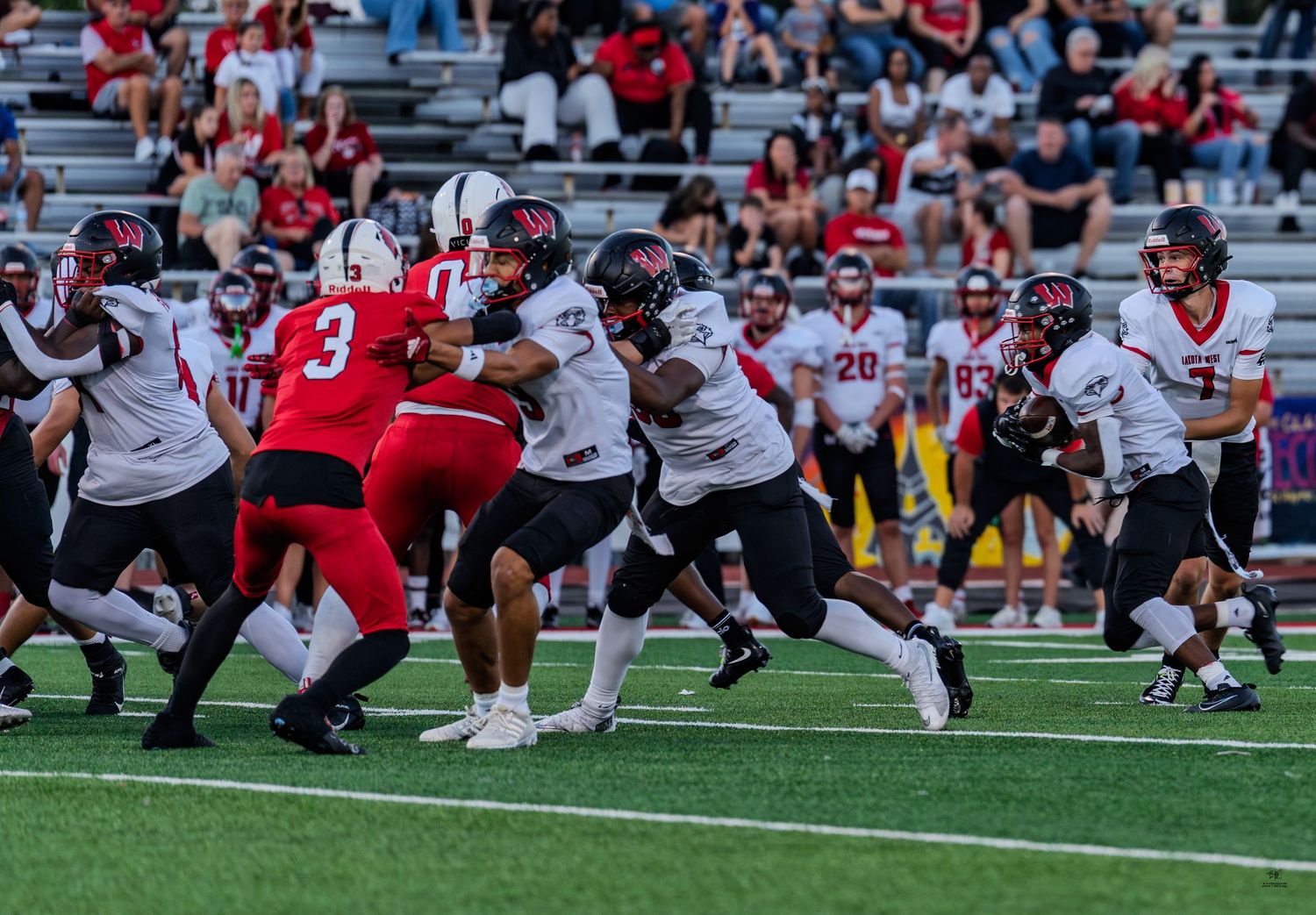 Football players in white uniforms push through the red team defensive line during a high school game.