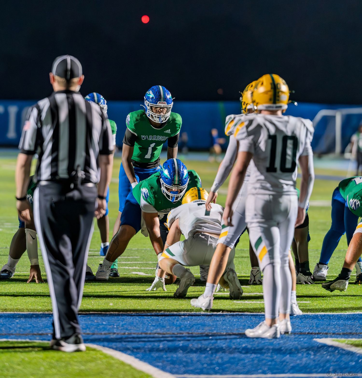 High school football teams face off at line of scrimmage during night game with referee watching.