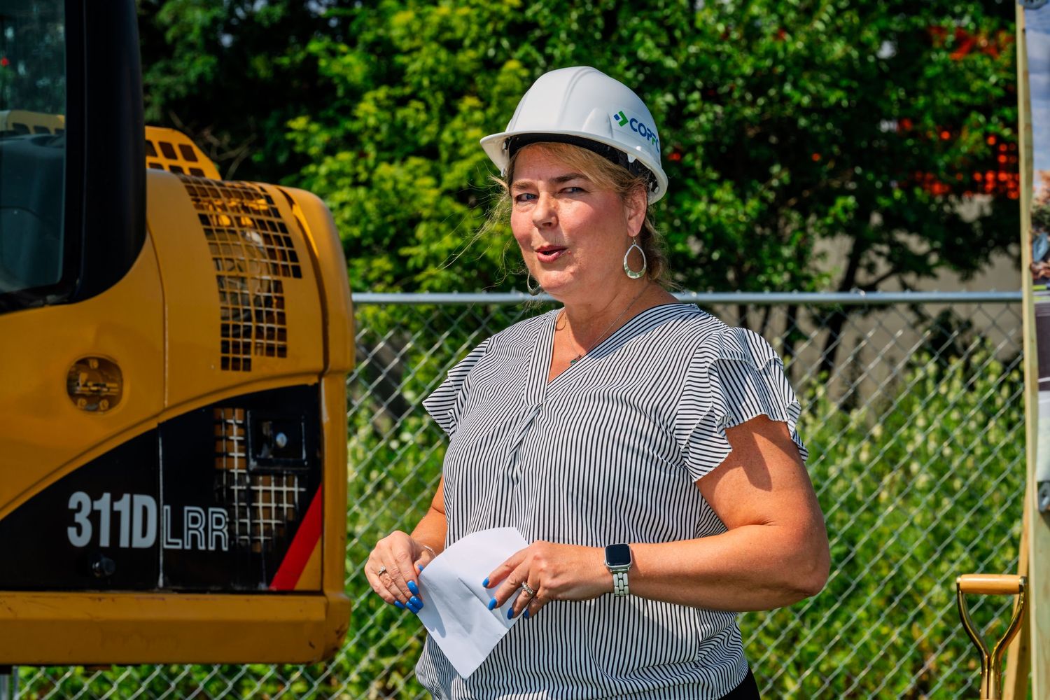 Woman in COPFCU hard hat speaking at a construction site near yellow equipment.