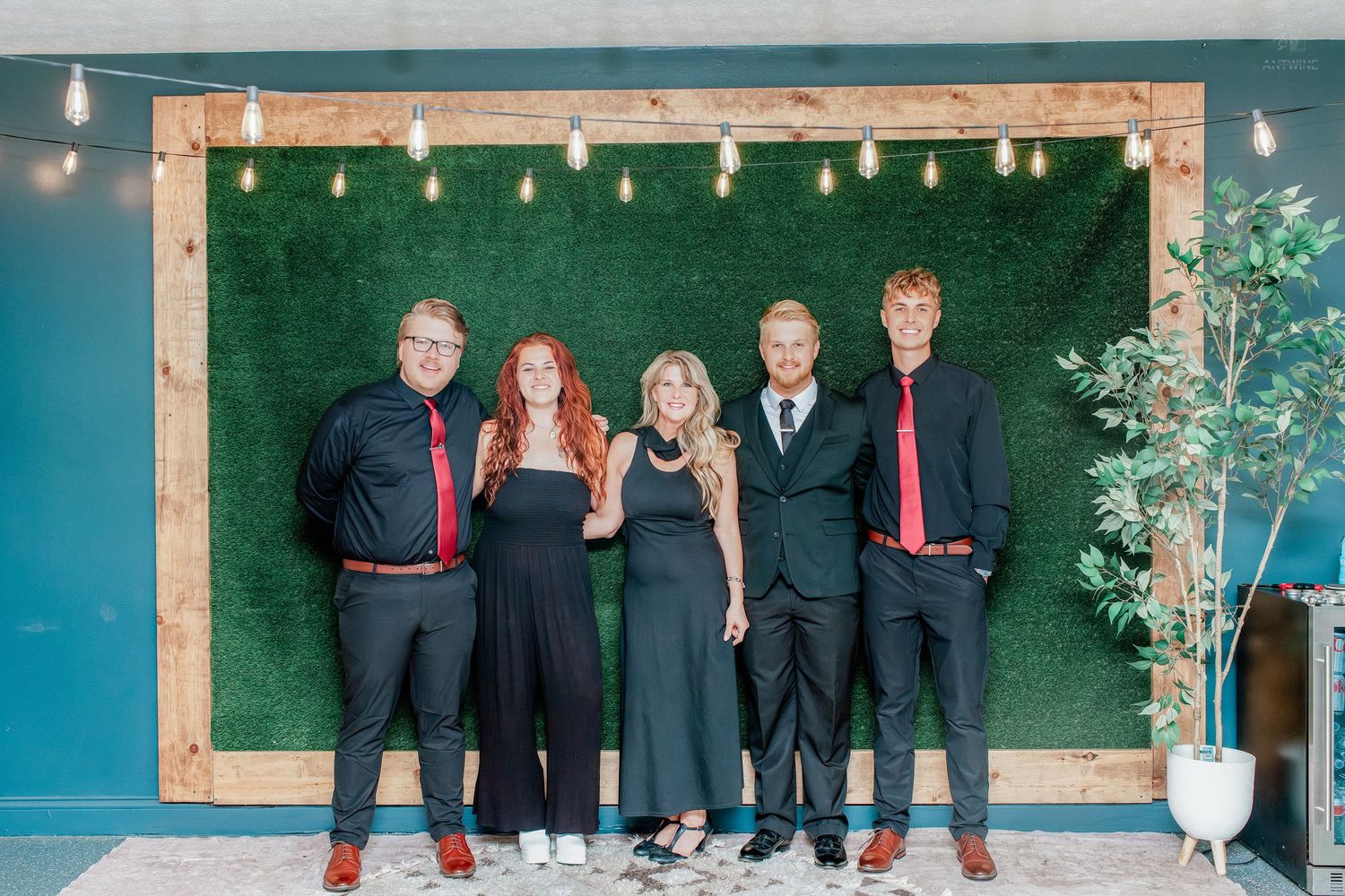 Five people in black formal attire pose smiling before a green hedge wall with string lights at an elegant event.