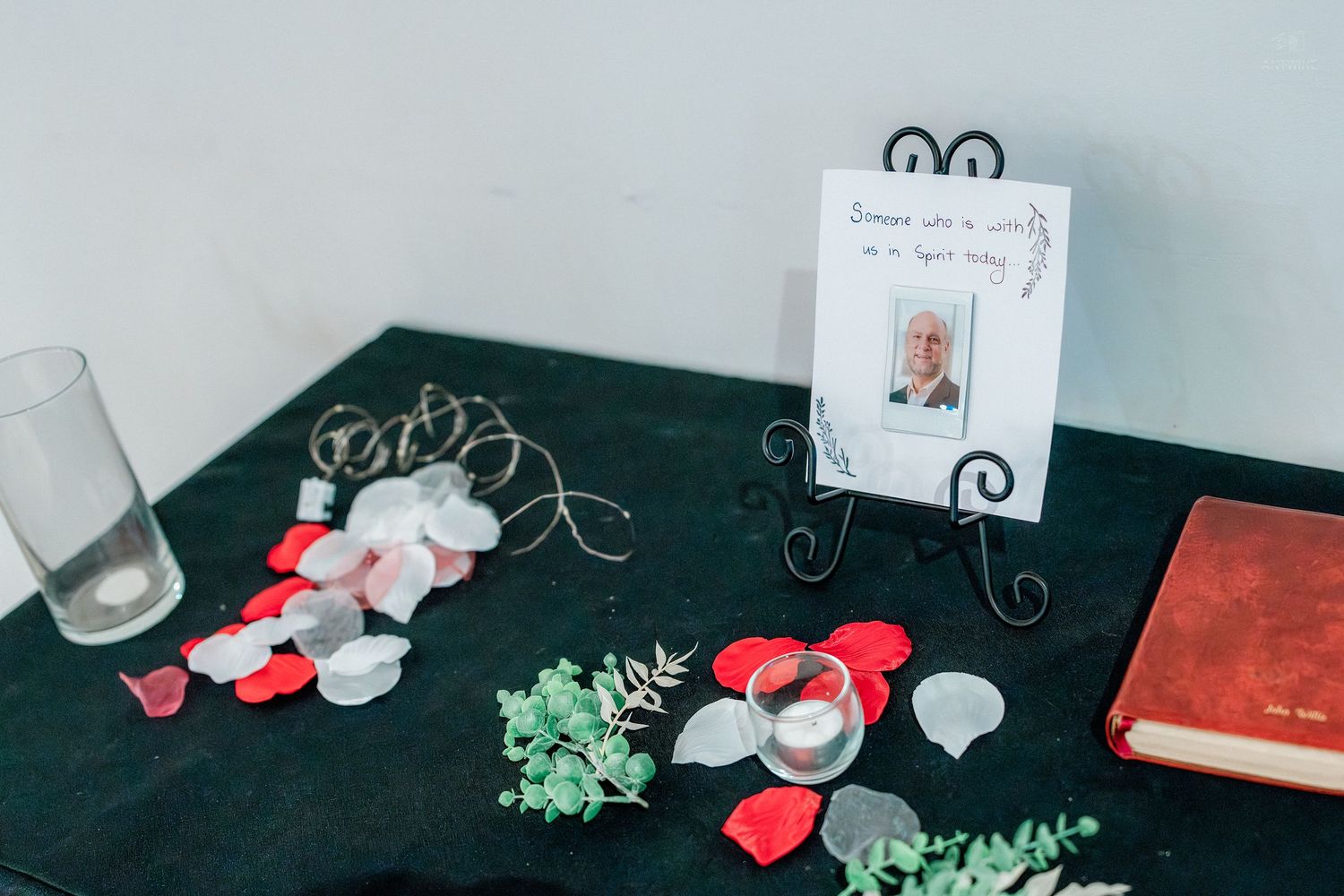 Memorial table with red and white rose petals, framed photo, candles, greenery, and a red book on black cloth.