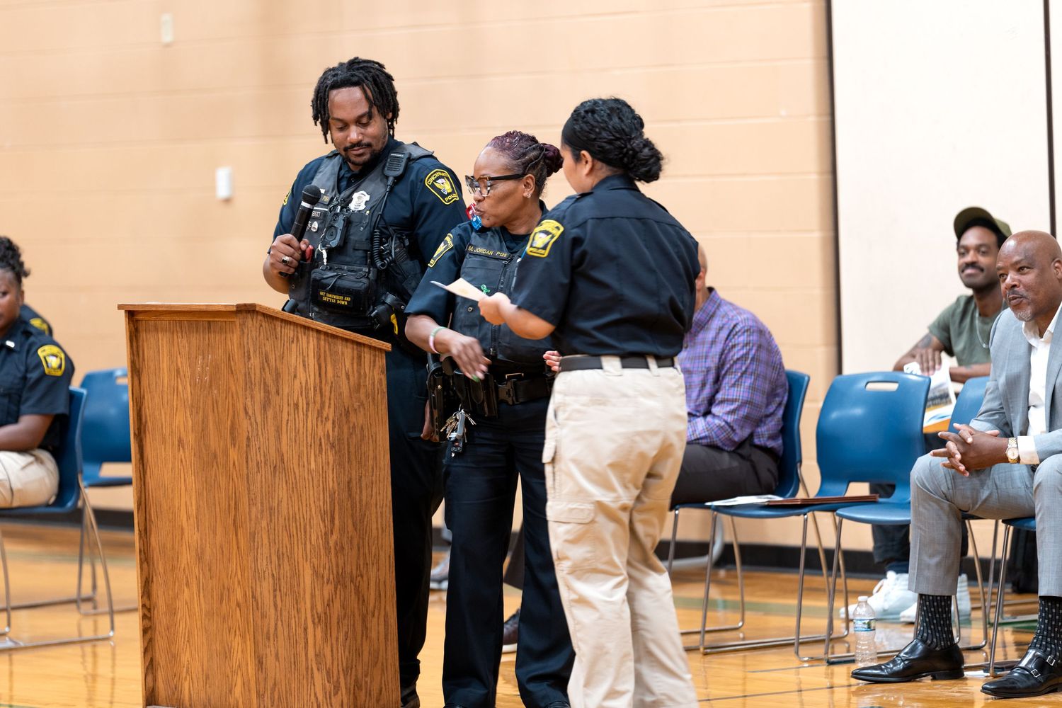 Police officers in uniform gather at a podium during a community law enforcement event inside a school gymnasium.