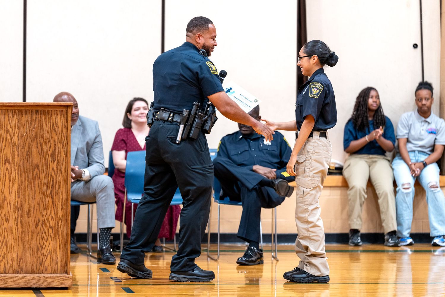 Police officer pins badge on graduate during law enforcement ceremony in school gymnasium.