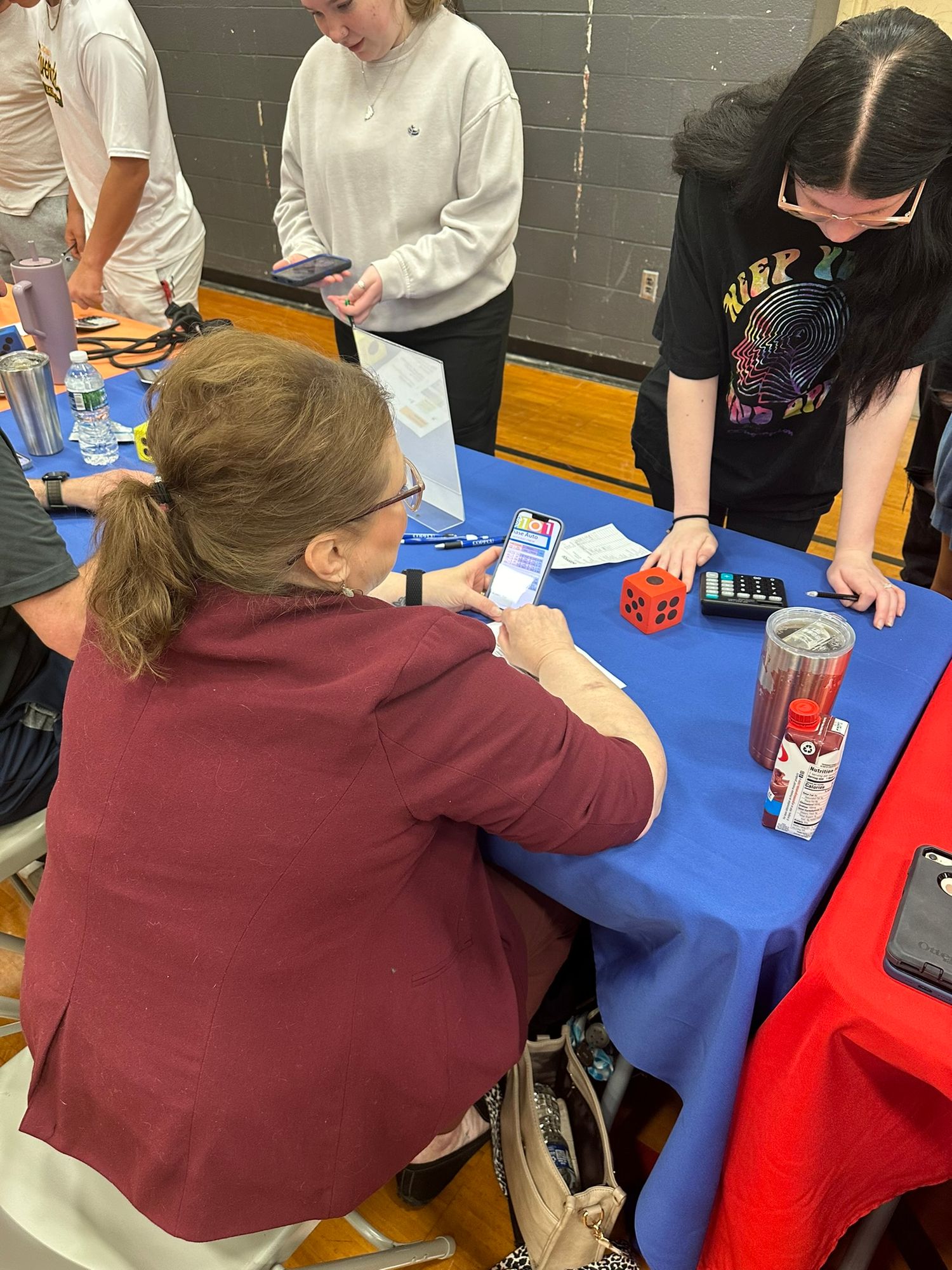 Students and teacher interact at a blue table with dice and phones during a classroom activity in a school gymnasium.