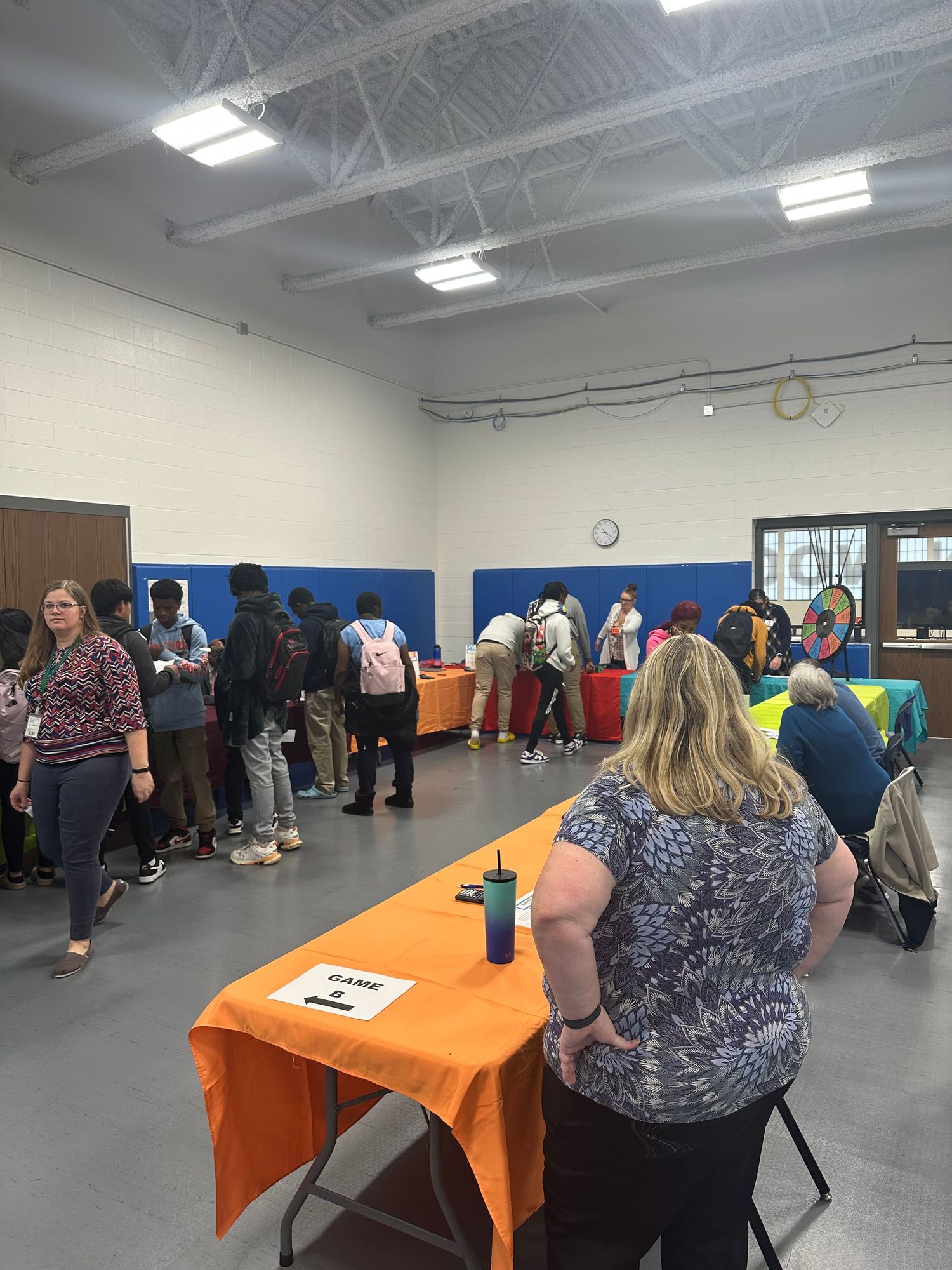 Students and teachers gather at colorful booths during a school fair event held in a gymnasium.
