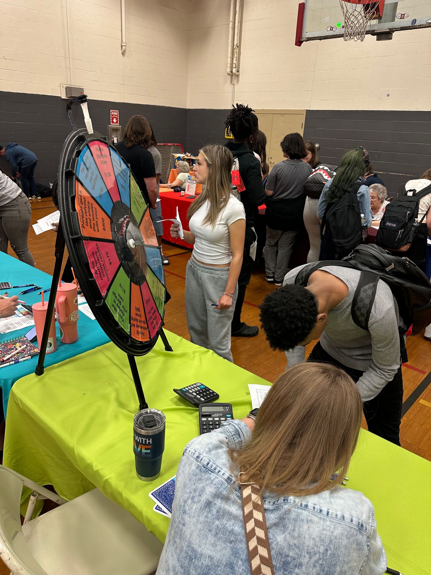 Students explore a colorful prize wheel at a school fair booth set up on a lime green table in a gym.