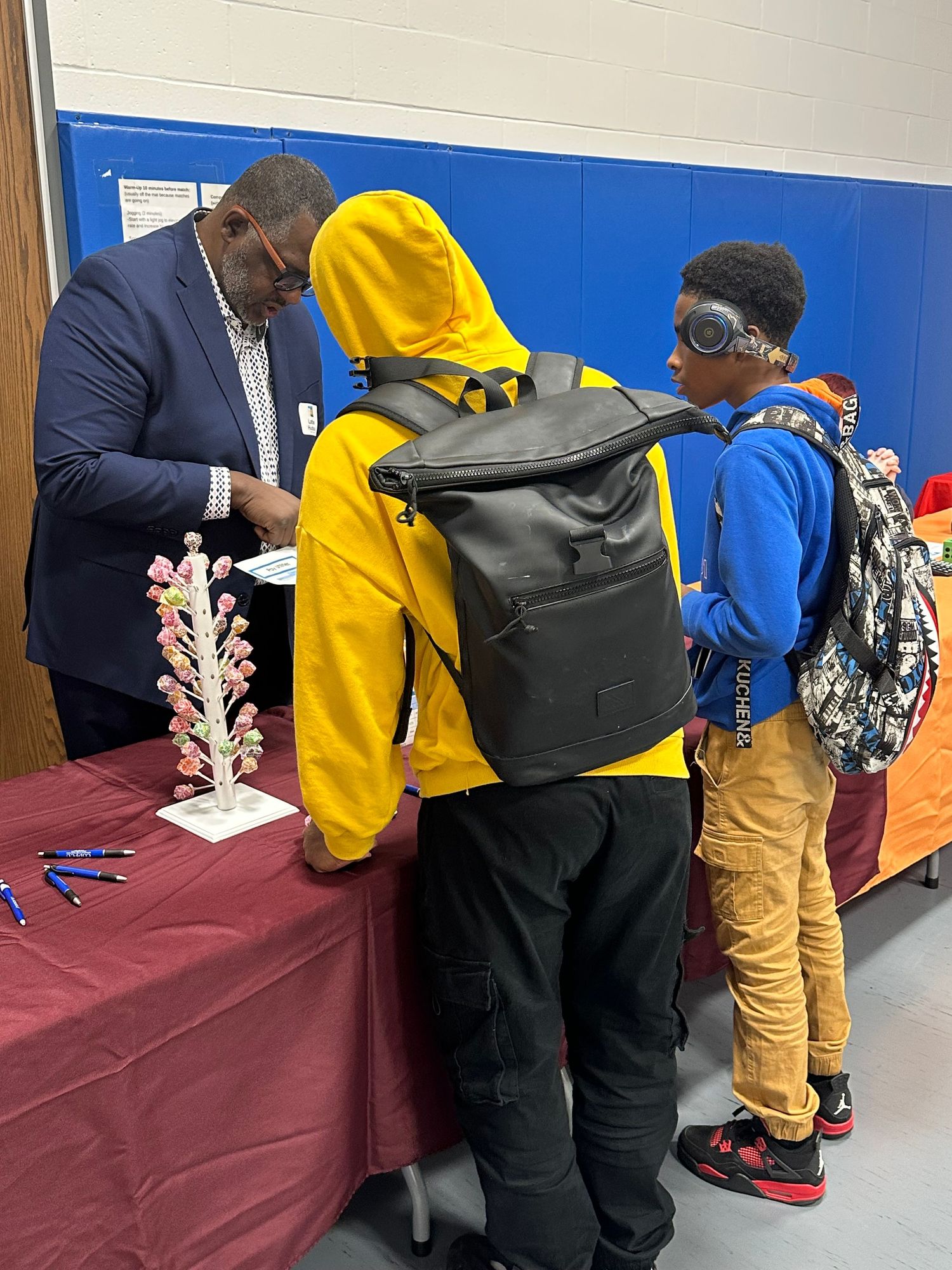 Students with backpacks interact with an adult presenter at an educational fair booth inside a school gymnasium.