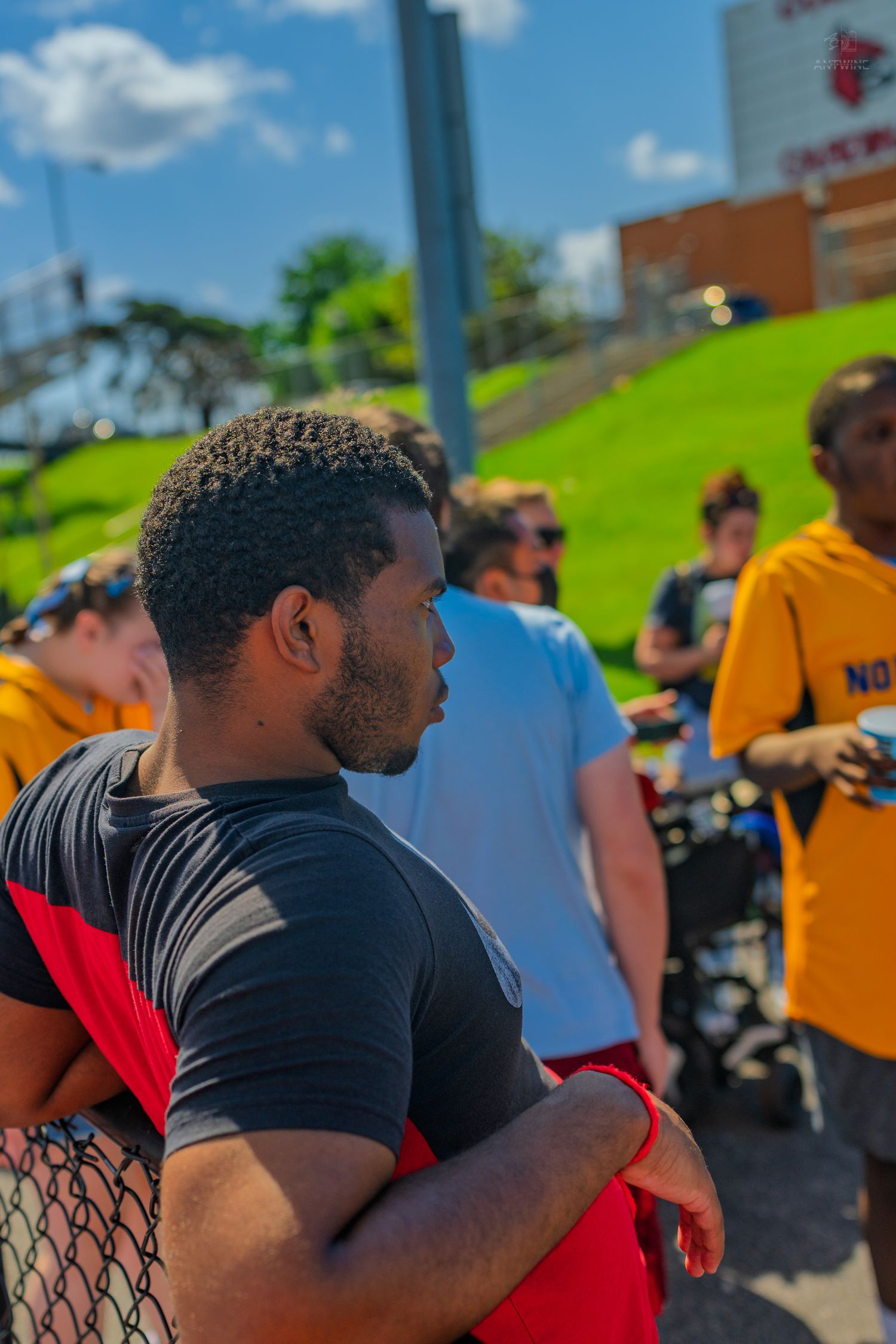 Young man in black and red shirt stands arms crossed at an outdoor gathering on a sunny day.