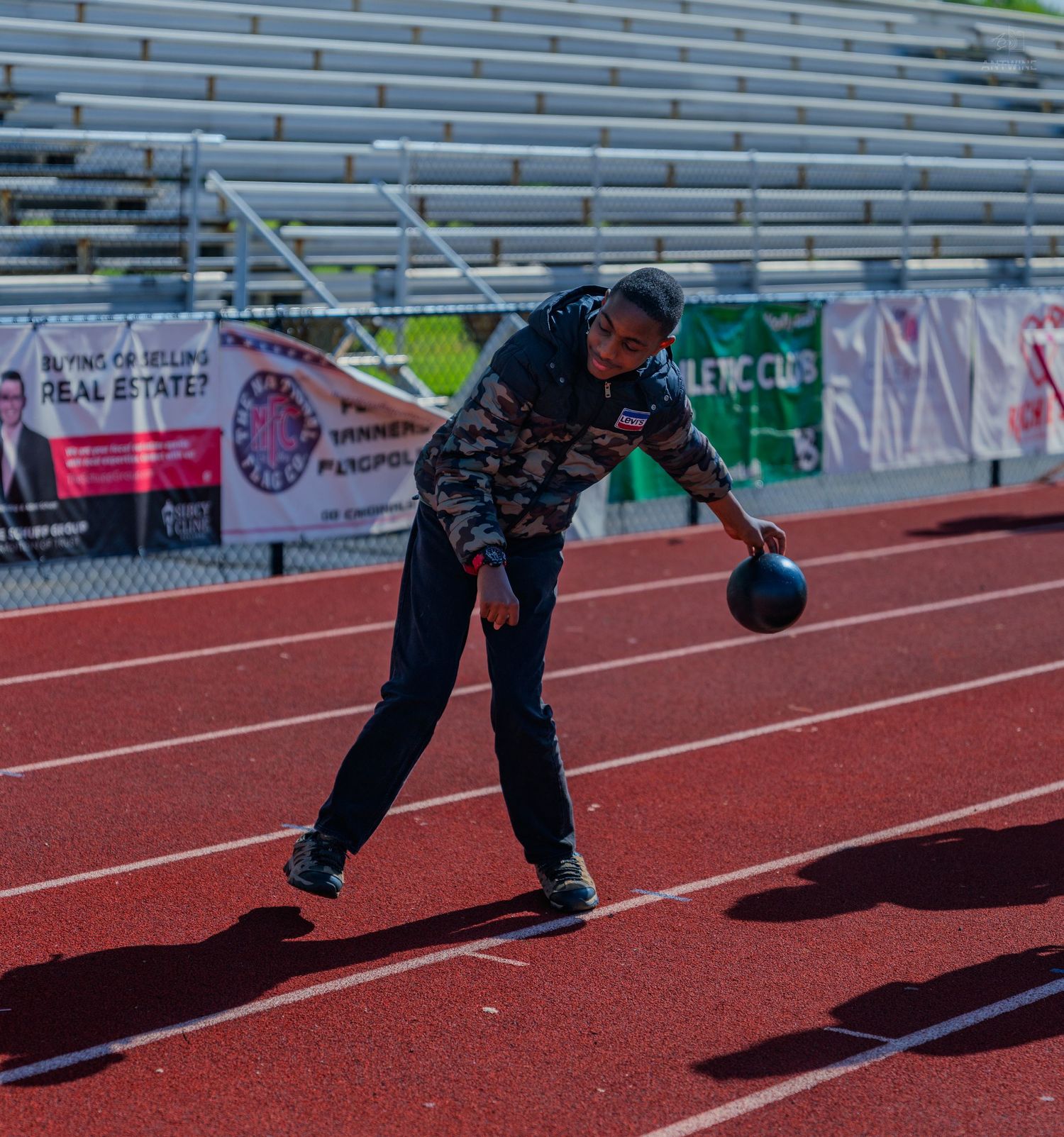 Athlete in camouflage jacket prepares to throw a bocce ball on a red track at an outdoor stadium.
