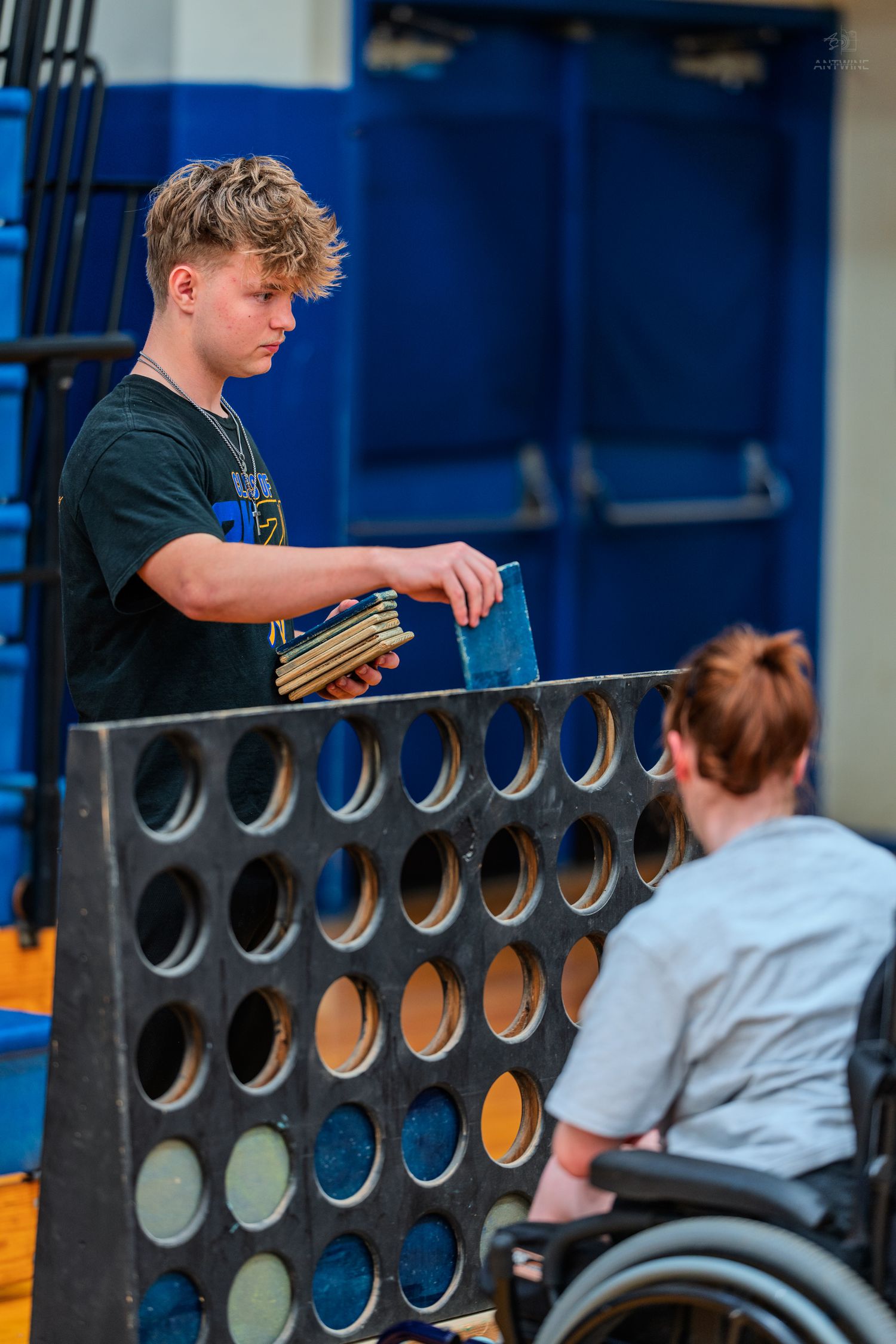 Teen boy plays giant Connect Four game in a gym while a person in a wheelchair watches nearby.