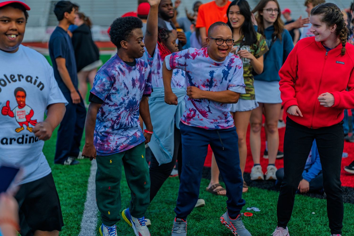 Students in tie-dye shirts dance and celebrate together on a sports field during a school event.