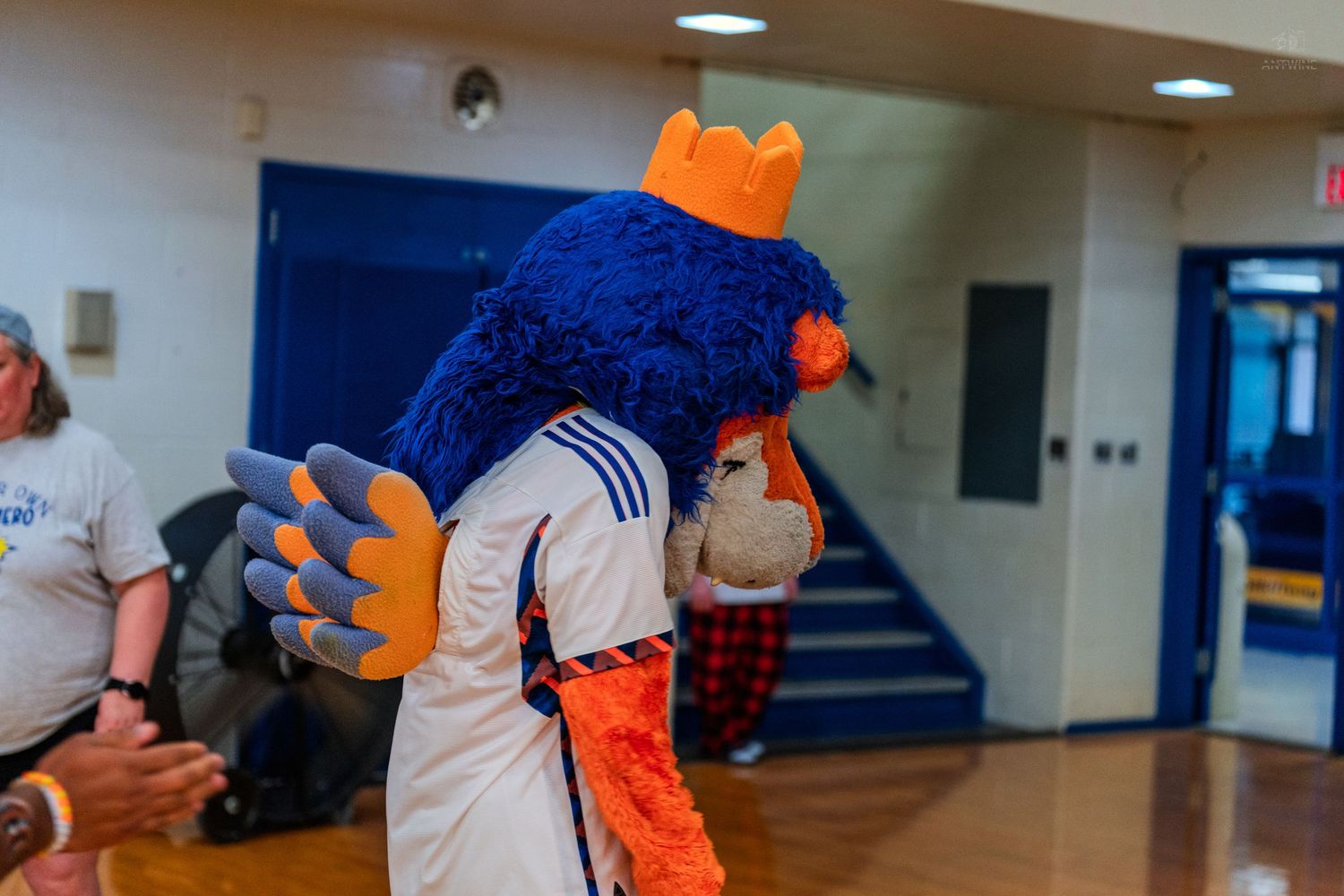 Blue fuzzy mascot with orange crown and claws wearing a white jersey at an indoor gymnasium event.