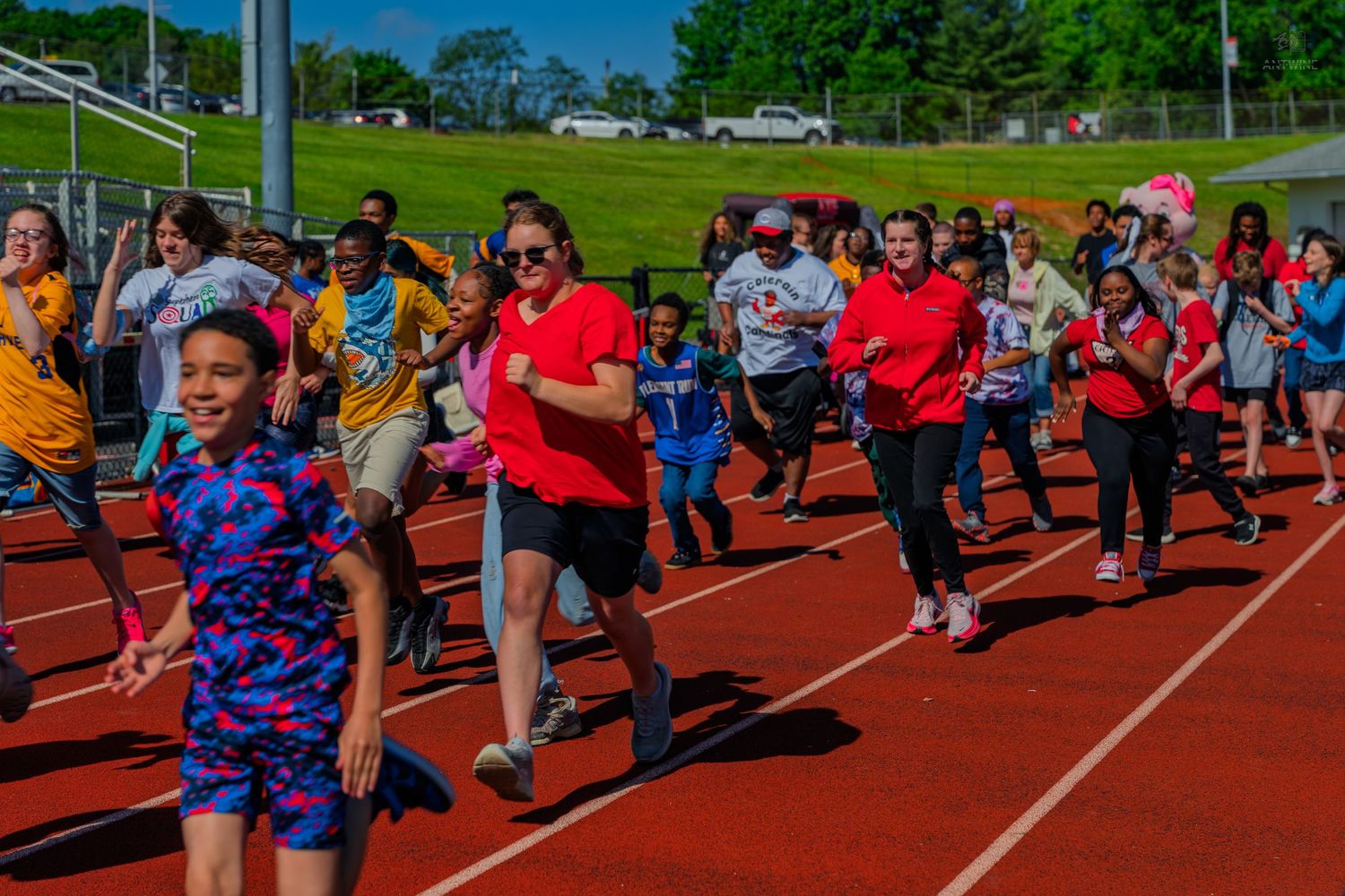 Children and adults running on a red track during an outdoor school field day event on a sunny spring day.