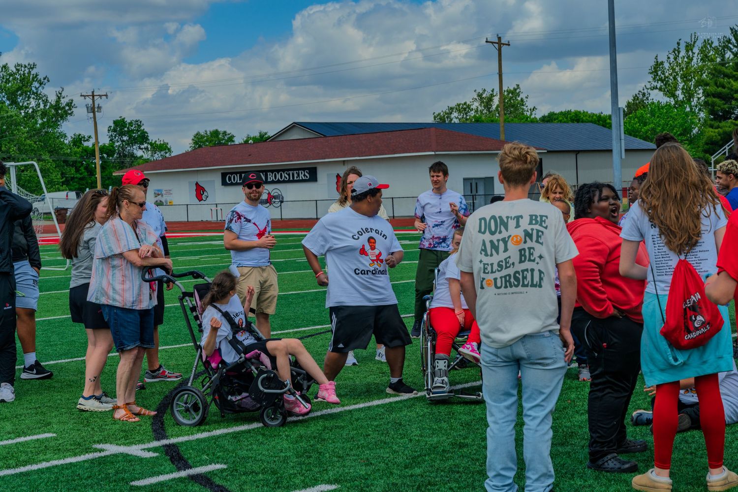 Group of people gathered on a green sports field during an outdoor event, with a school building visible in the background.
