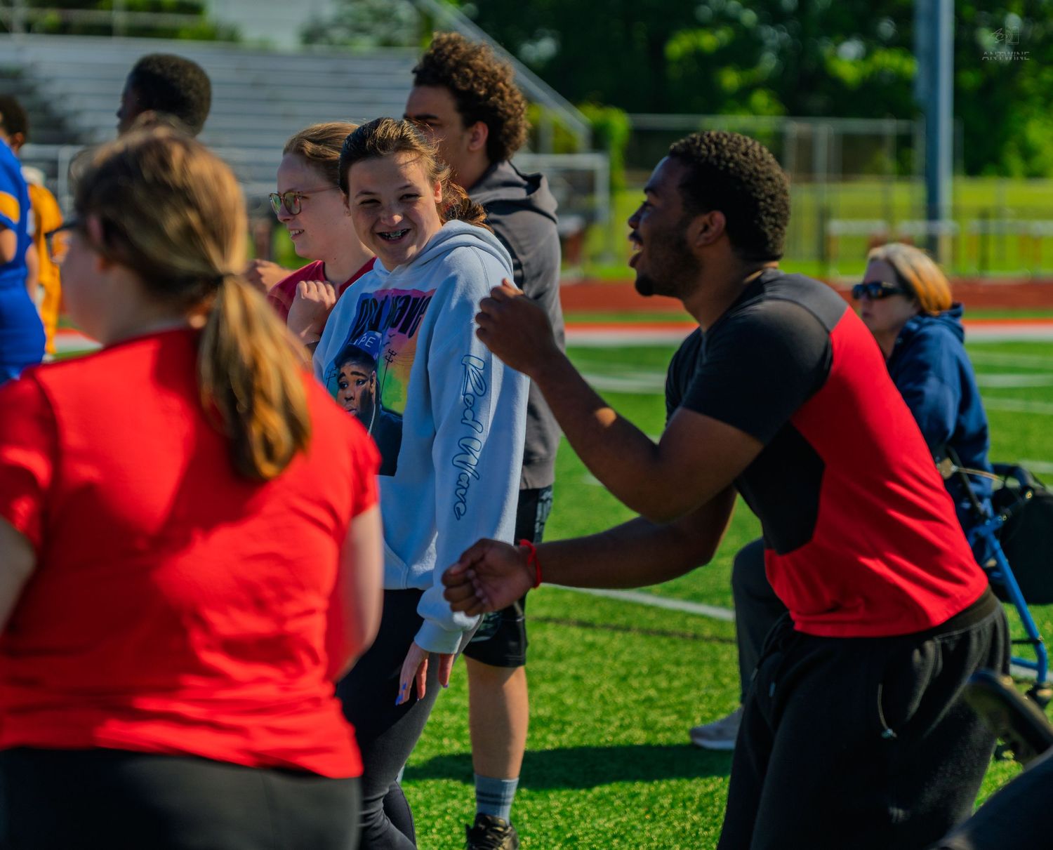 Athletes and coaches interact warmly on a green sports field during an outdoor athletic event on a sunny day.