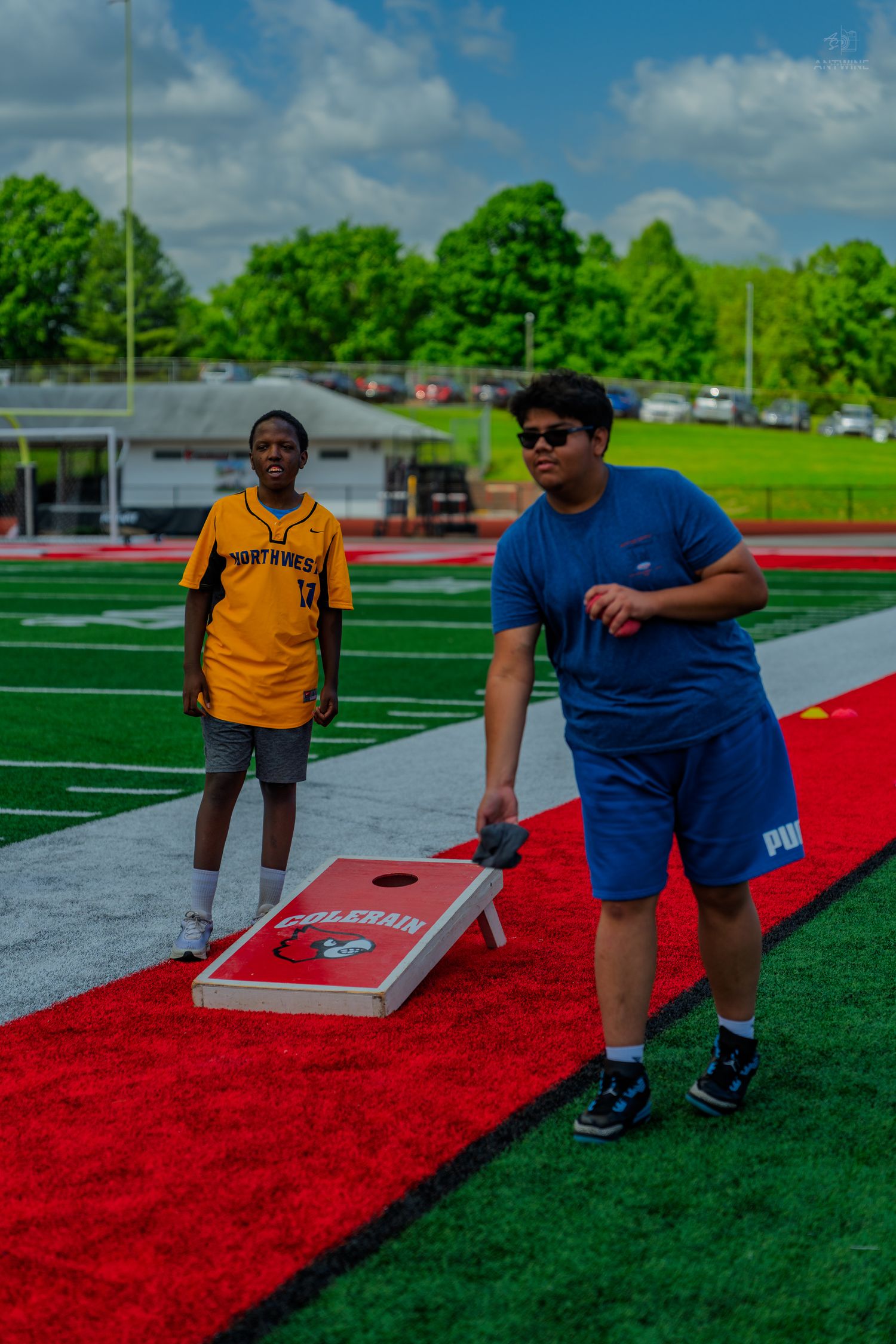 Two young men playing cornhole on a red and green turf football field on a cloudy day.
