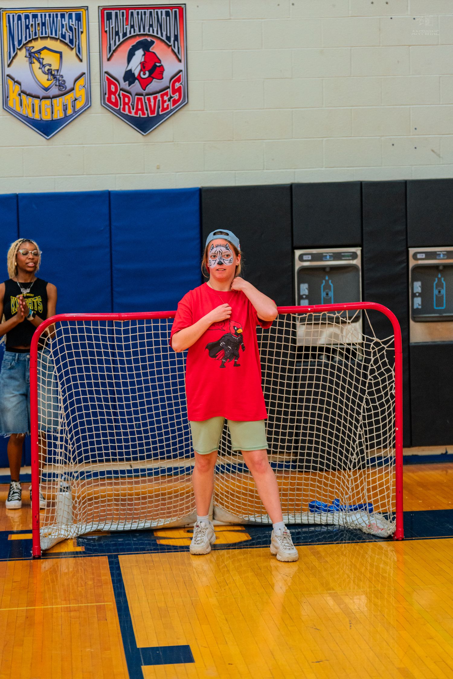 Student in red shirt stands in front of hockey goal in school gymnasium with team banners on wall.