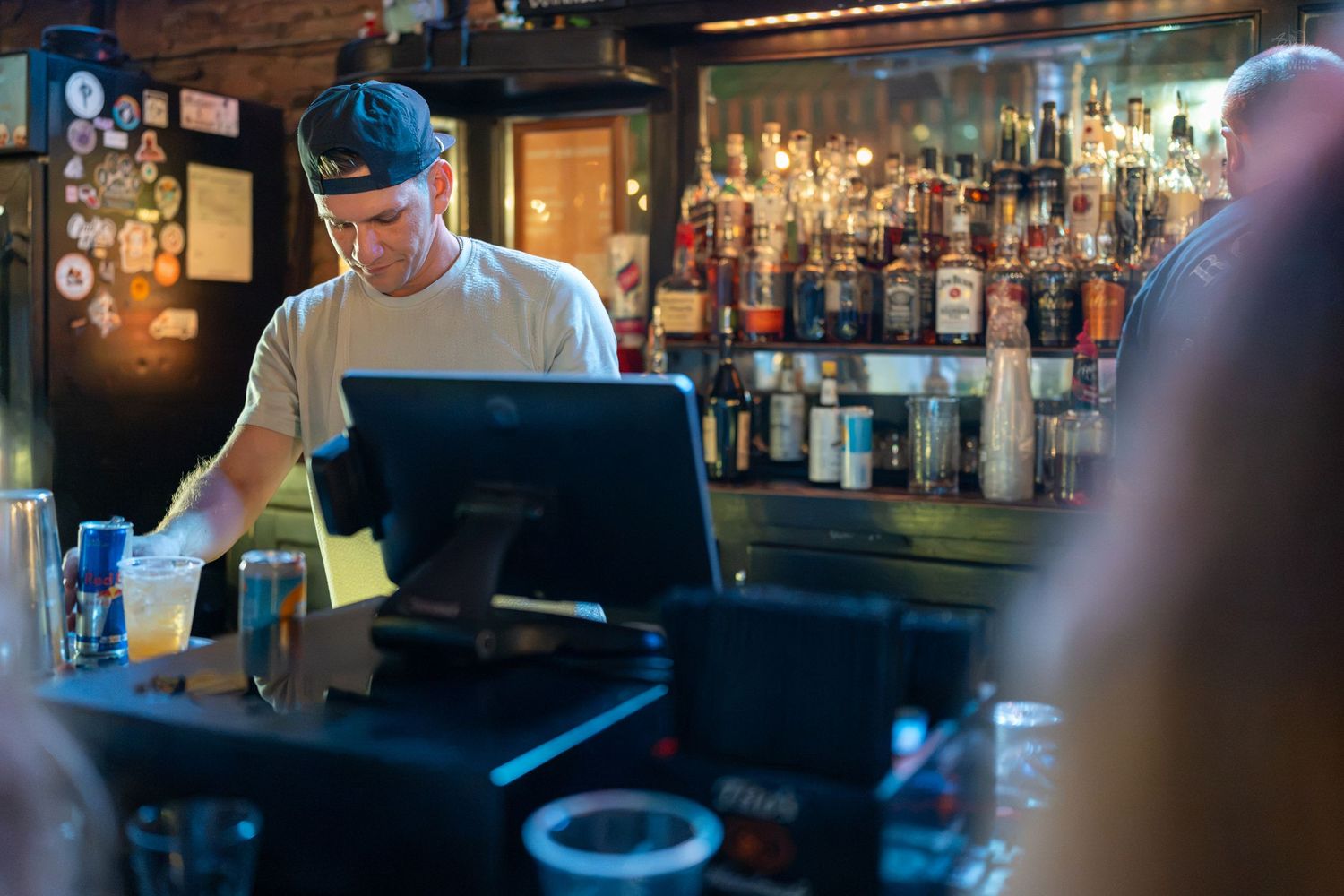 Bartender working behind a bar counter with a POS system, surrounded by shelves of liquor bottles in a dimly lit bar.