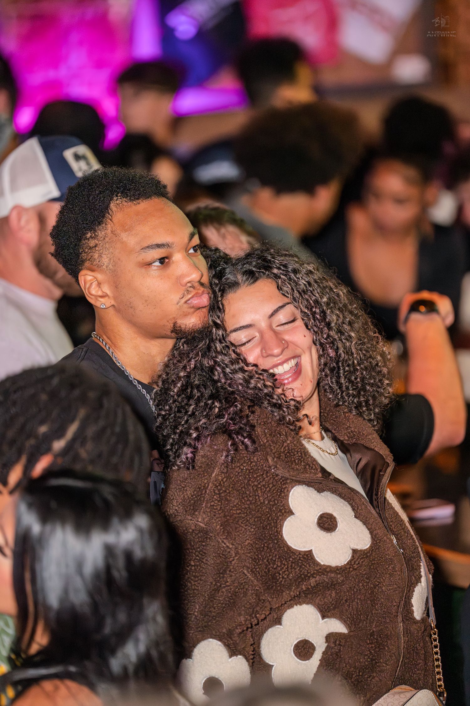 Young couple embracing and smiling at a lively indoor bar event with colorful lighting.