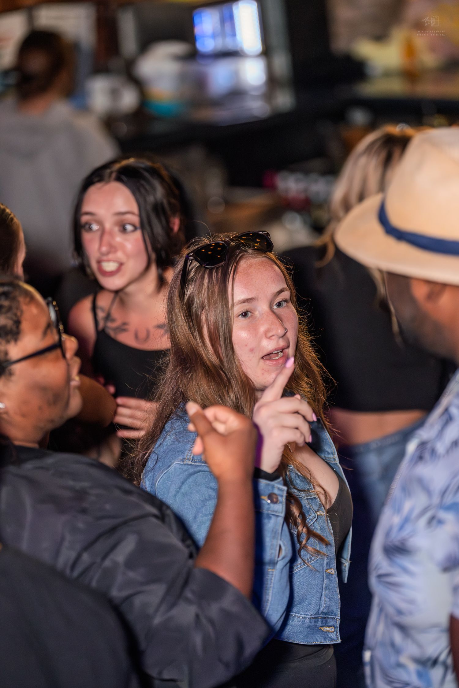 Young woman in denim jacket engages in conversation at a busy bar with friends gathered around her.