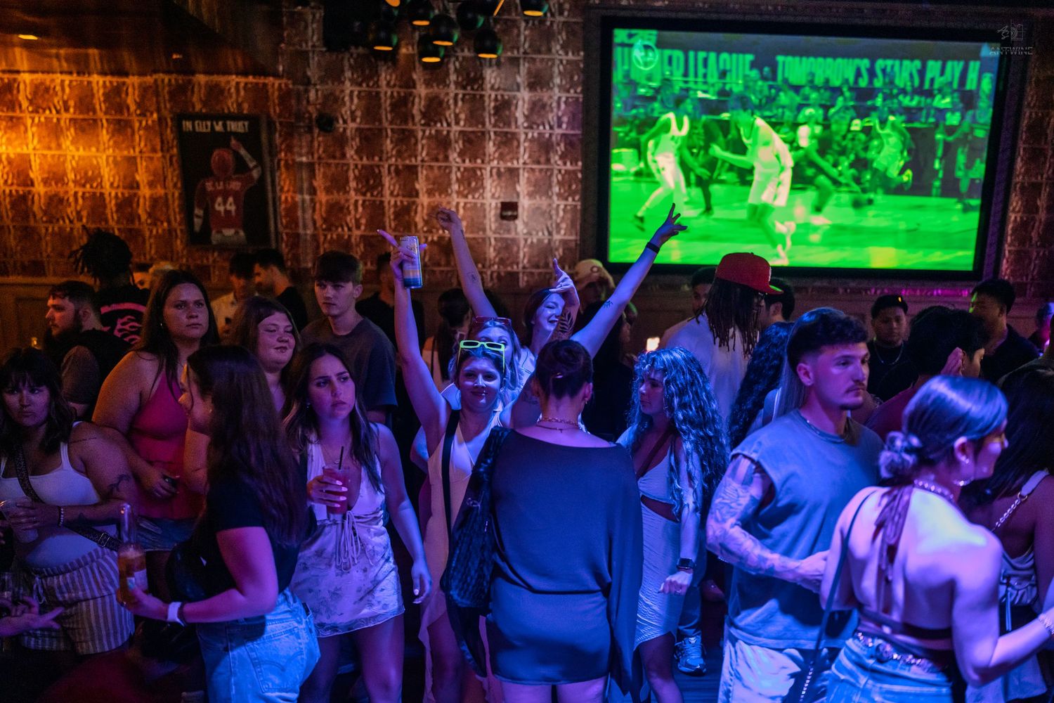 Energetic nightclub crowd cheering with hands raised as a sports game plays on a large screen.