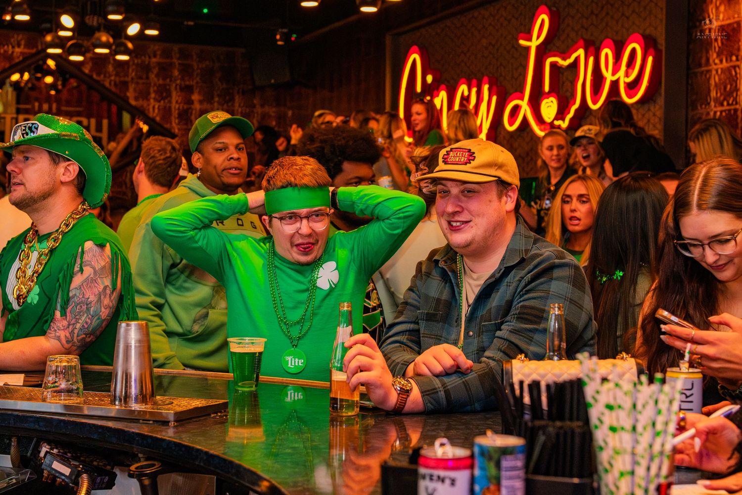 Colorful St. Patricks Day bar scene with people in green outfits under a City Love neon sign.