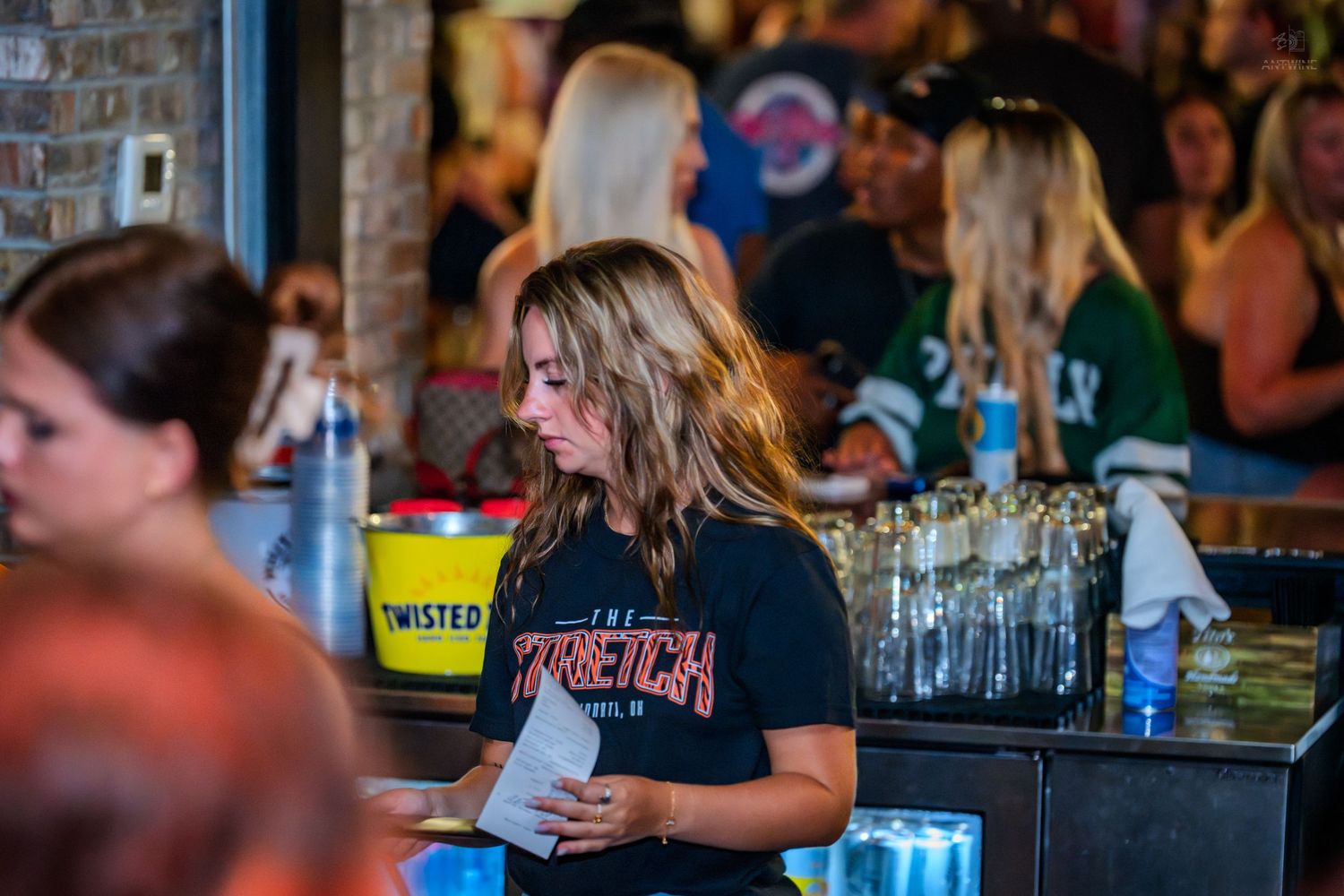 Female bartender in black shirt working behind a busy bar counter with glasses and drinks.