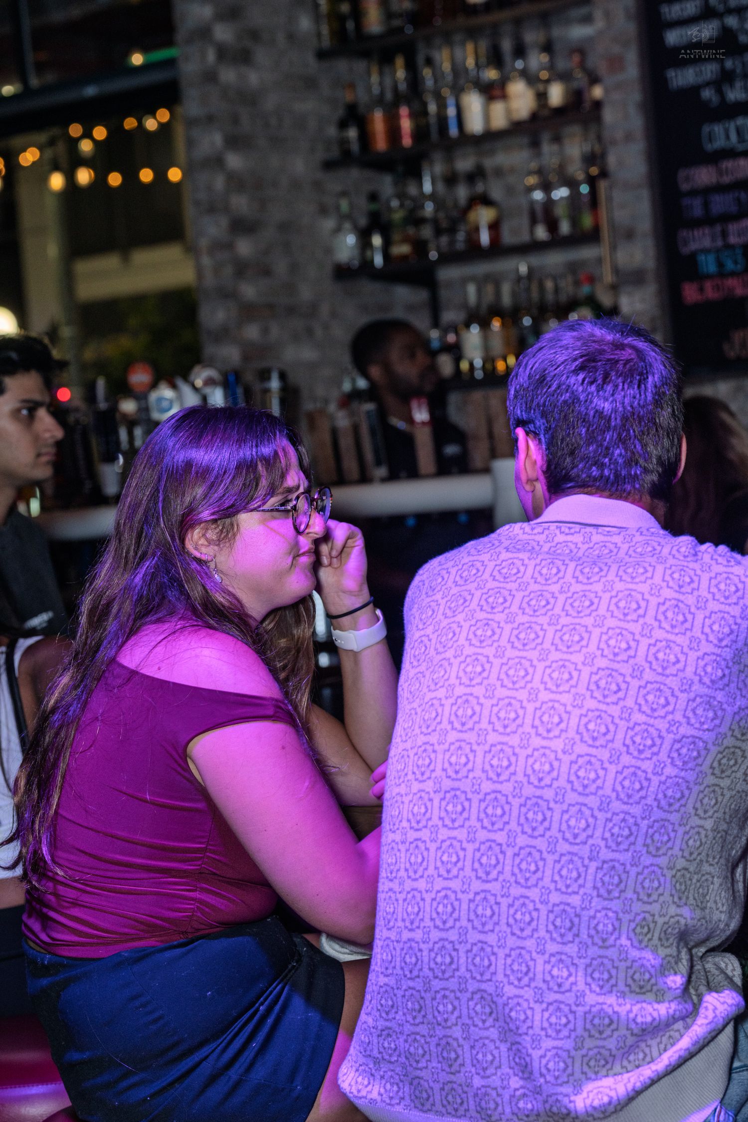 Woman with glasses in burgundy top chatting with man in patterned shirt at a purple-lit bar.