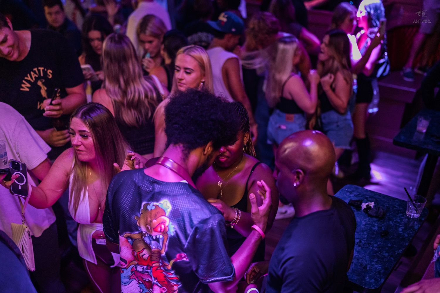 Diverse crowd of people socializing and dancing under purple lights at a nightclub party.