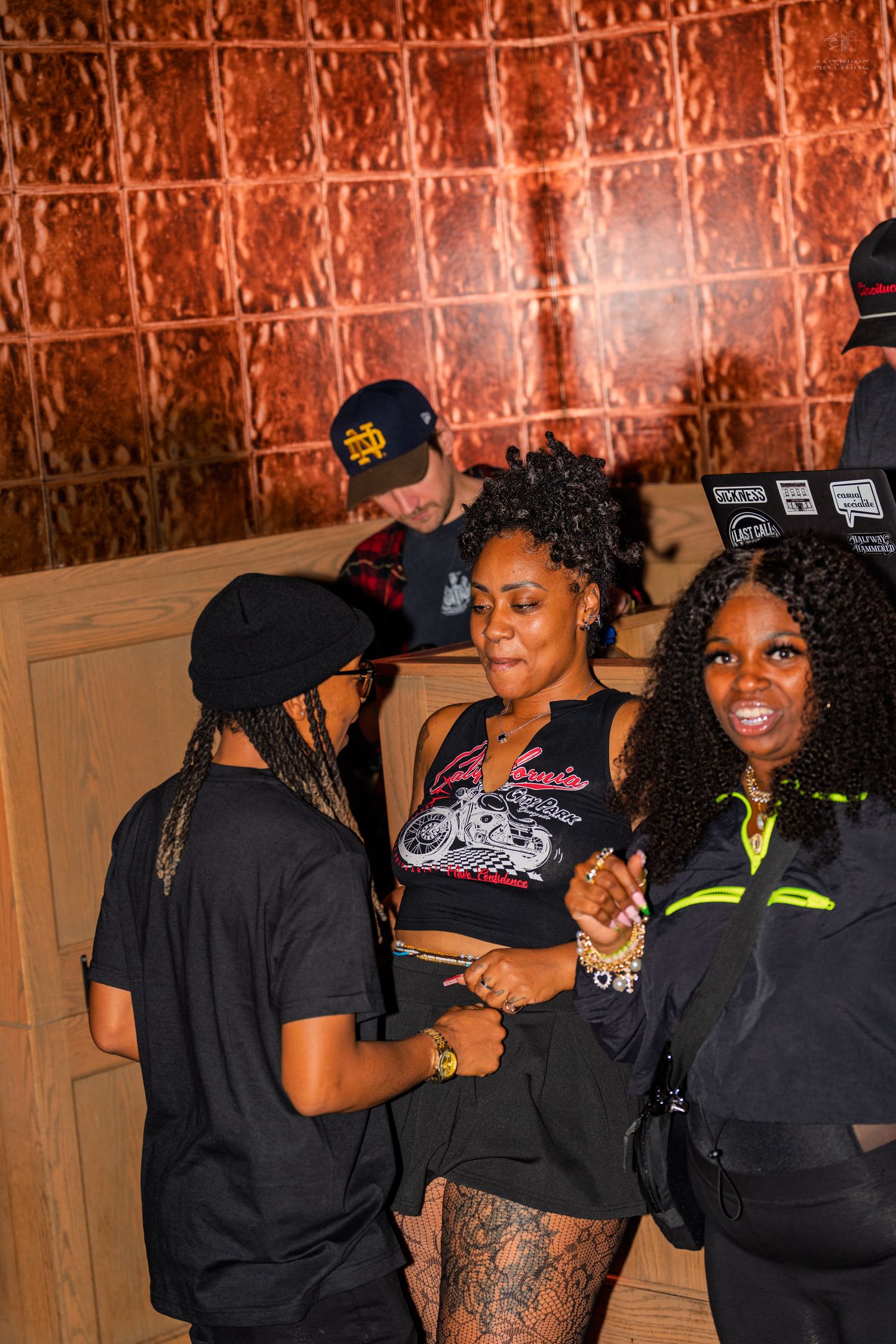 Three women in black outfits socializing near a DJ booth with copper-tiled walls.