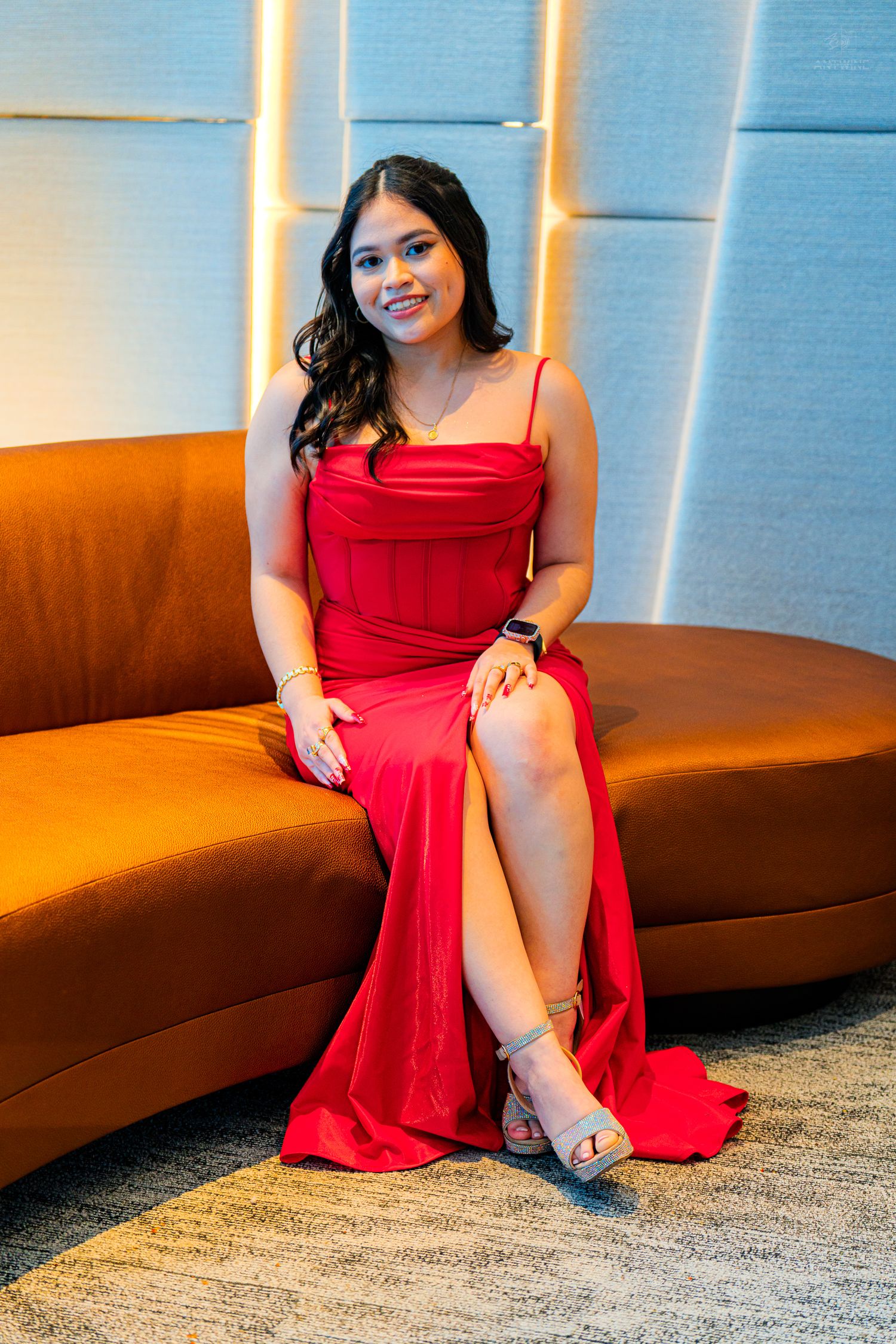 Young woman in elegant red satin gown with slit sitting on orange curved sofa at formal event.