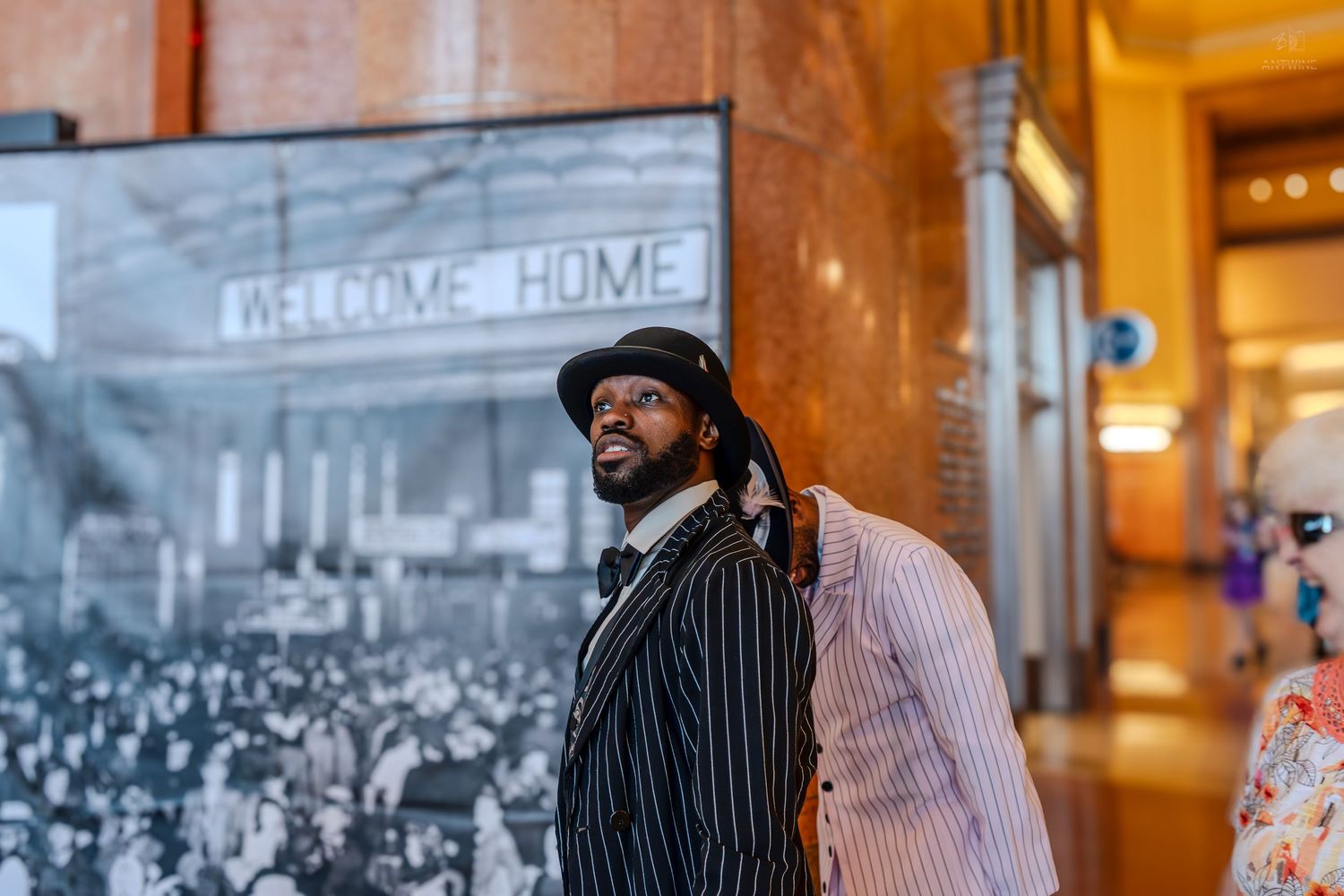 Man in pinstripe suit poses near a Welcome Home sign mural in a grand historic building interior.