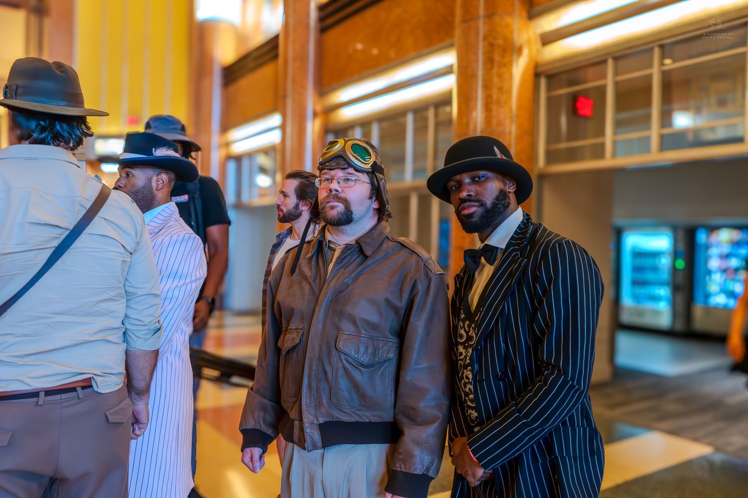 Men in vintage steampunk and zoot suit costumes socializing in a lobby at a themed event.
