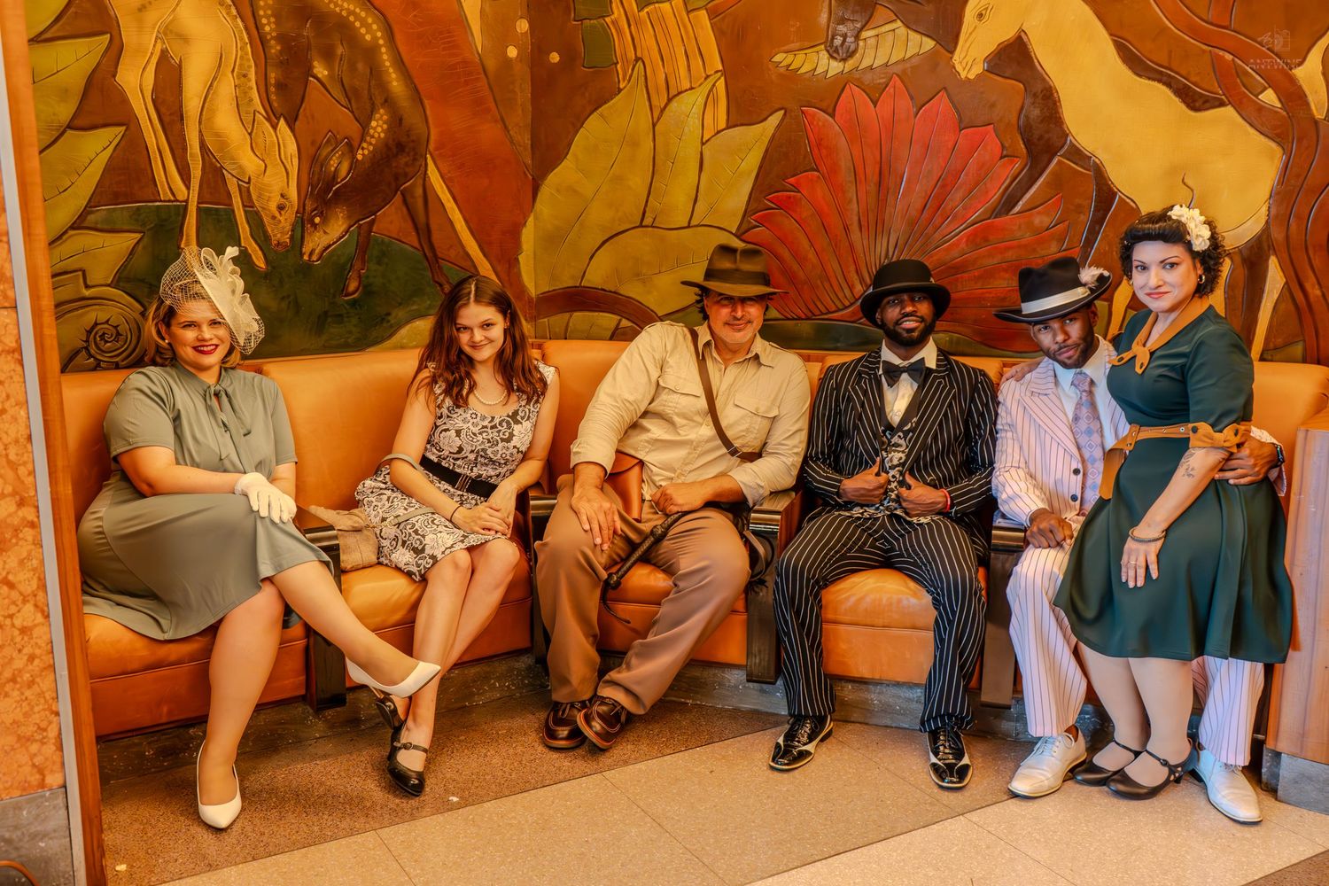 Six people in vintage 1940s fashion sitting on an orange sofa in front of a colorful Art Deco painted mural.