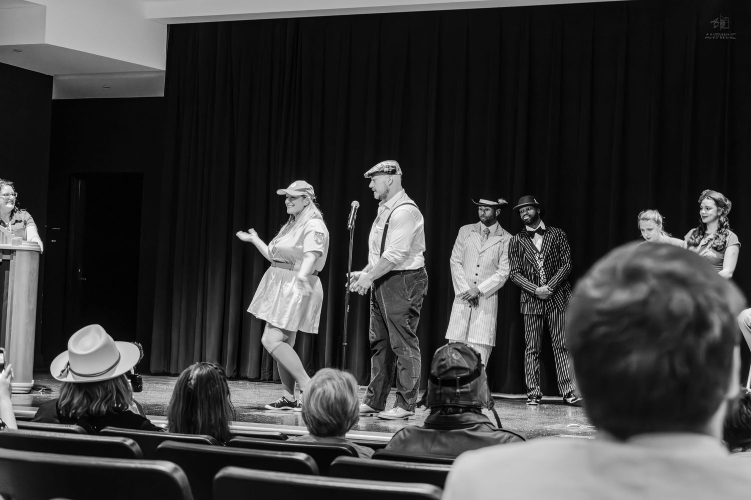 Black and white photo of performers on stage during a theater presentation with audience seated in rows.