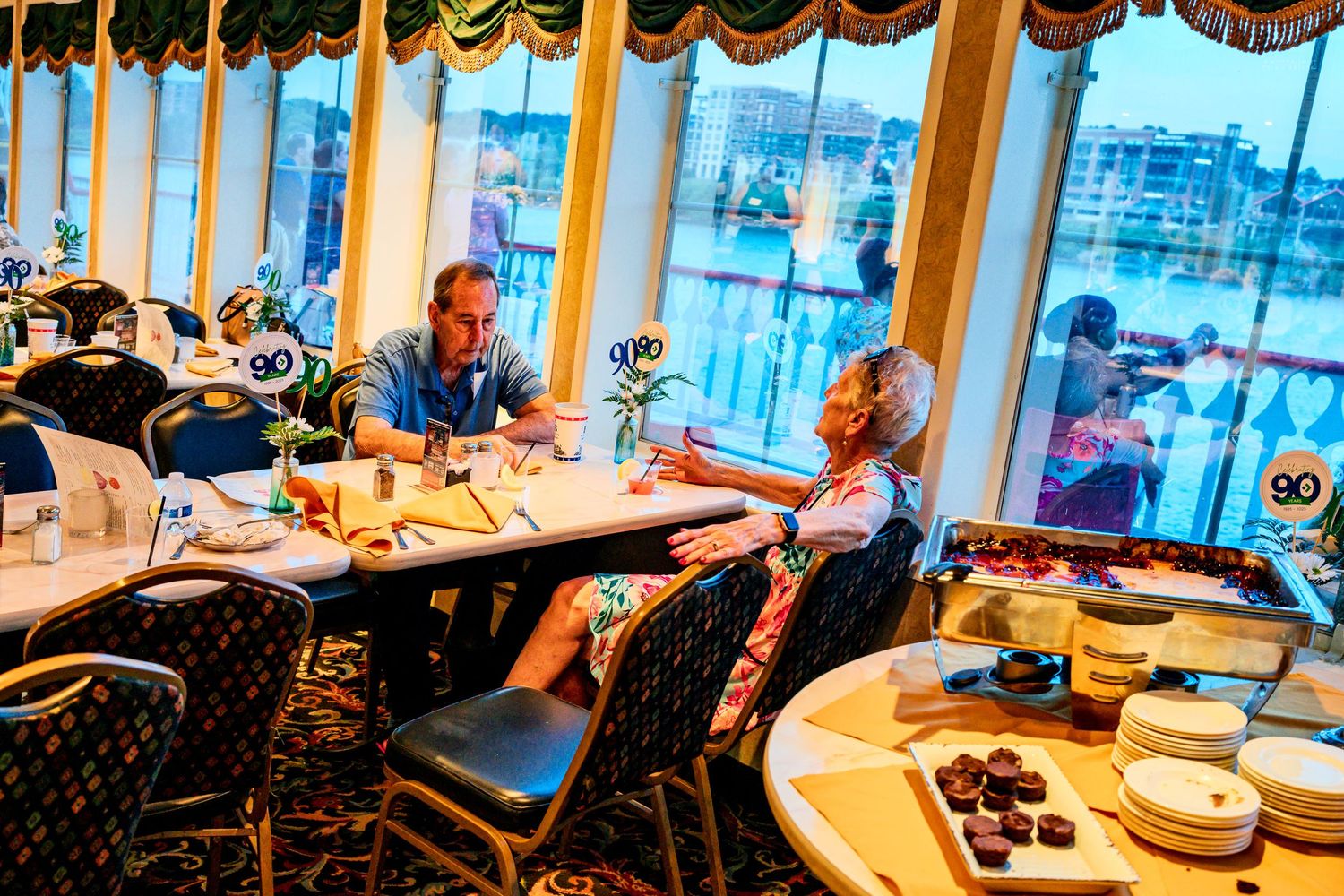 Couple dining at a cruise ship restaurant with panoramic windows overlooking a marina at dusk.