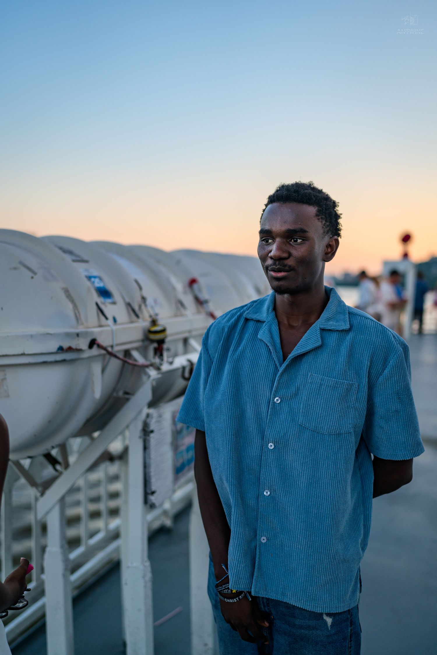 Young man in blue shirt stands on boat deck at dusk with equipment and water visible behind him.