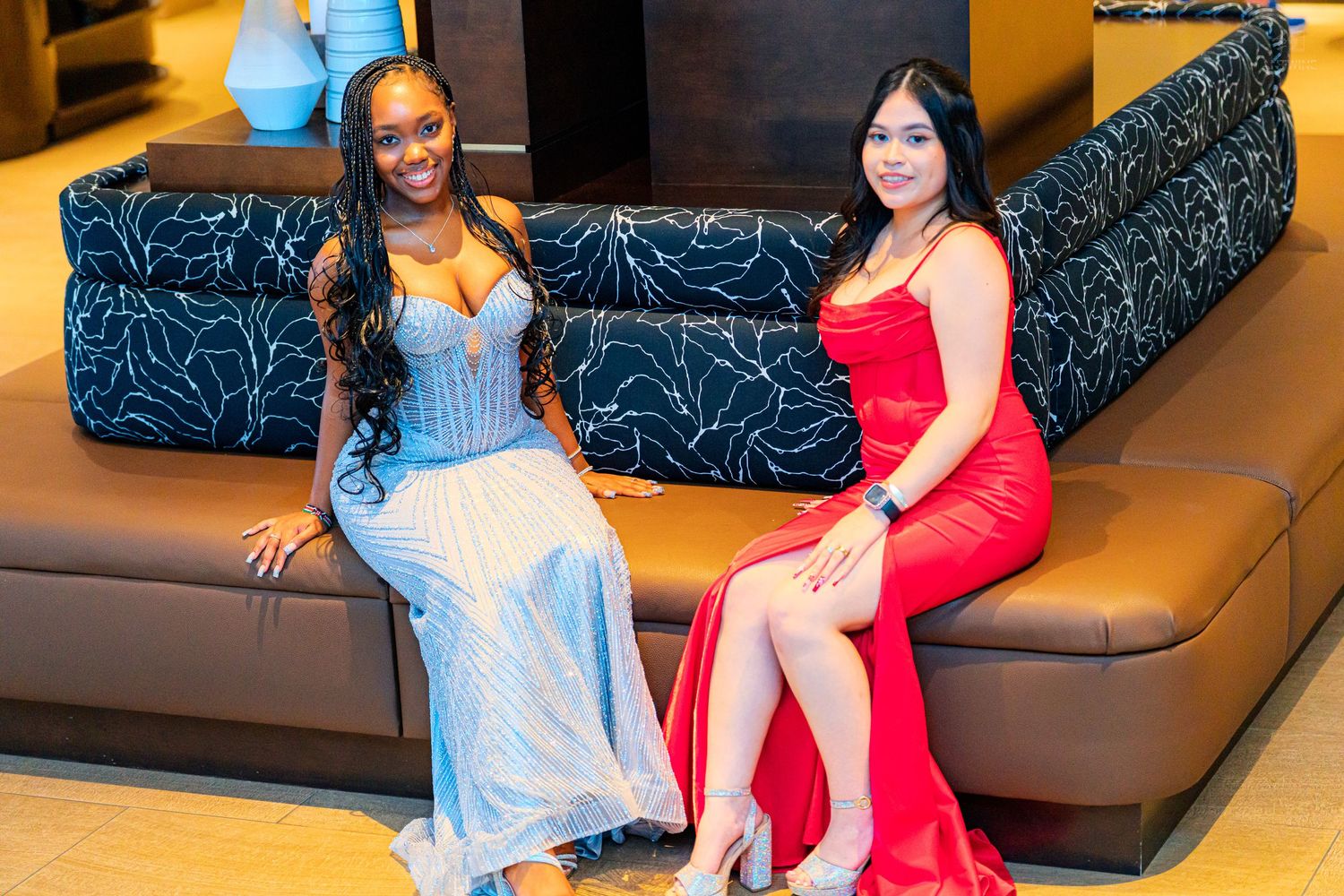 Two young women in formal gowns, one silver and one red, posing together on a leather sofa.