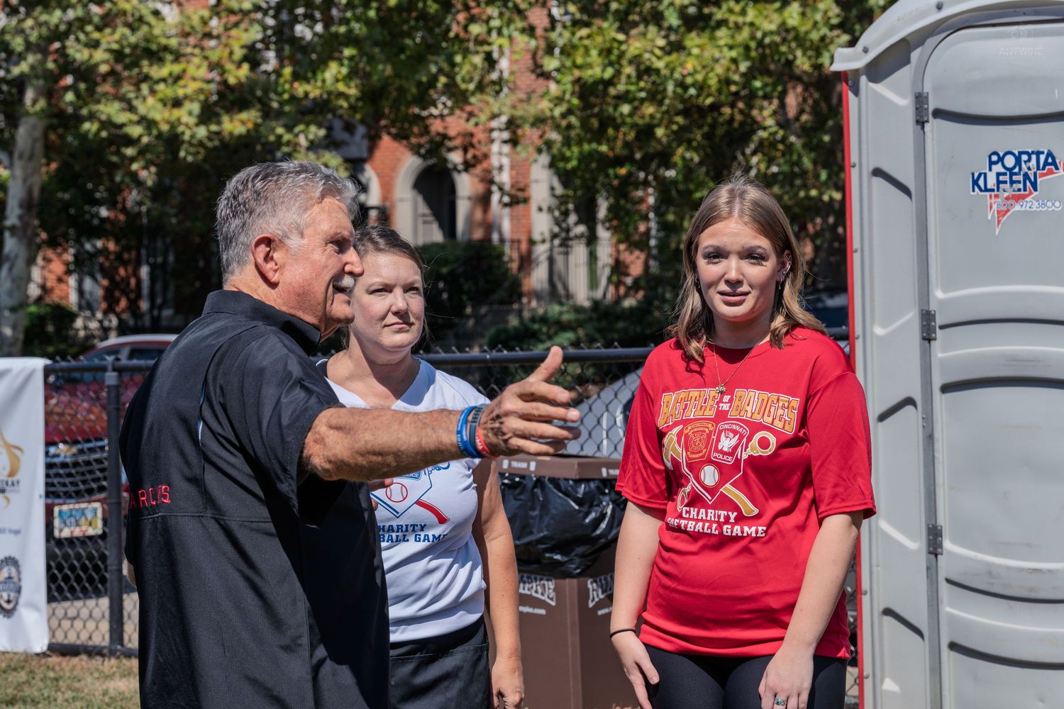 Man pointing while talking to two young women near a portable toilet at an outdoor sports event.