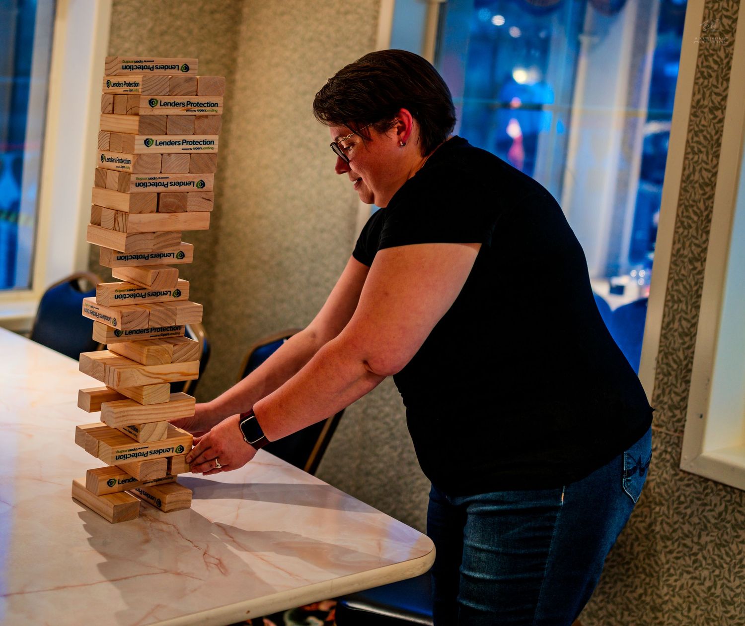 A person carefully removes a wooden block from a tall Jenga tower on a white table.