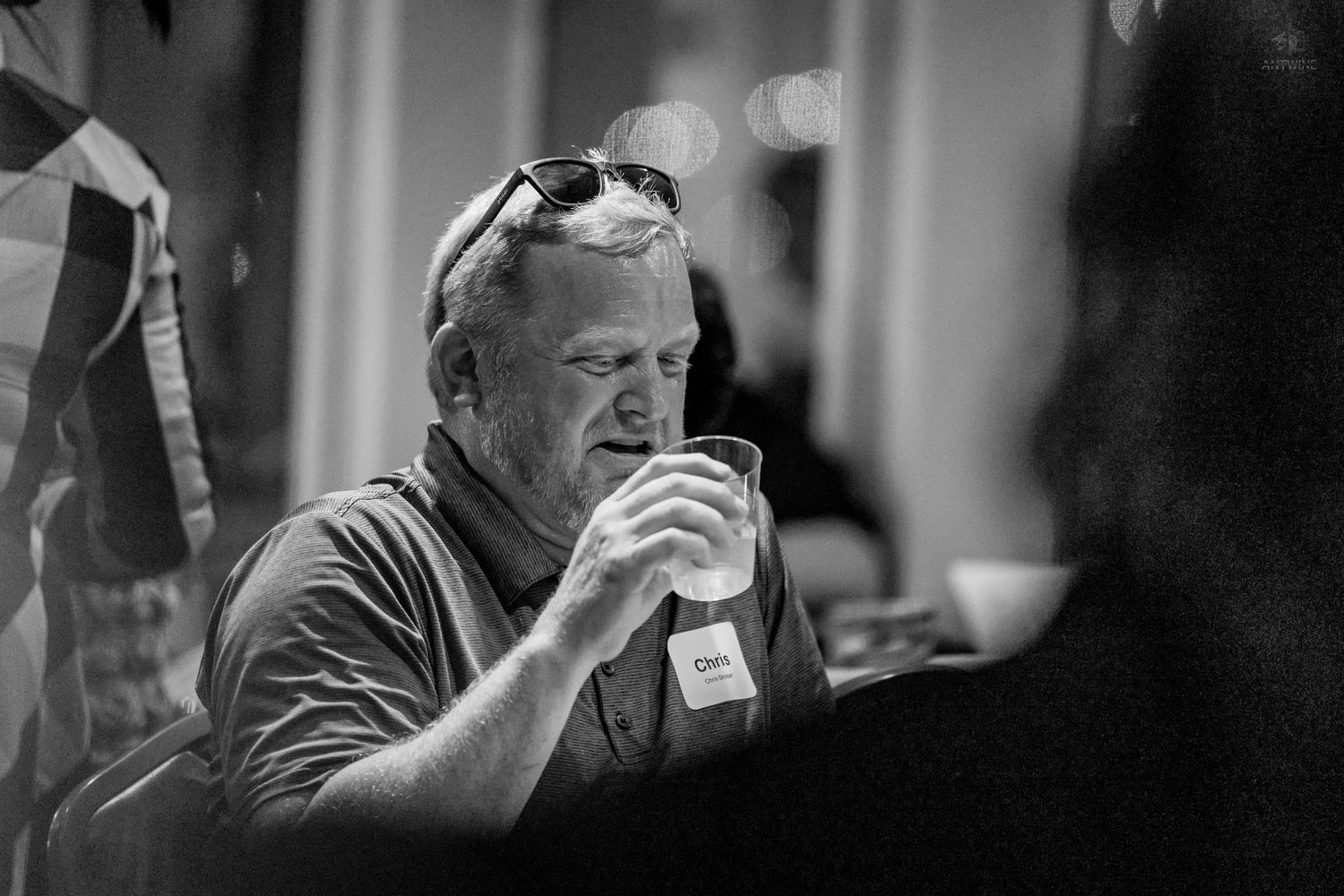 Black and white photo of a man holding a drink at a casual social gathering or event.