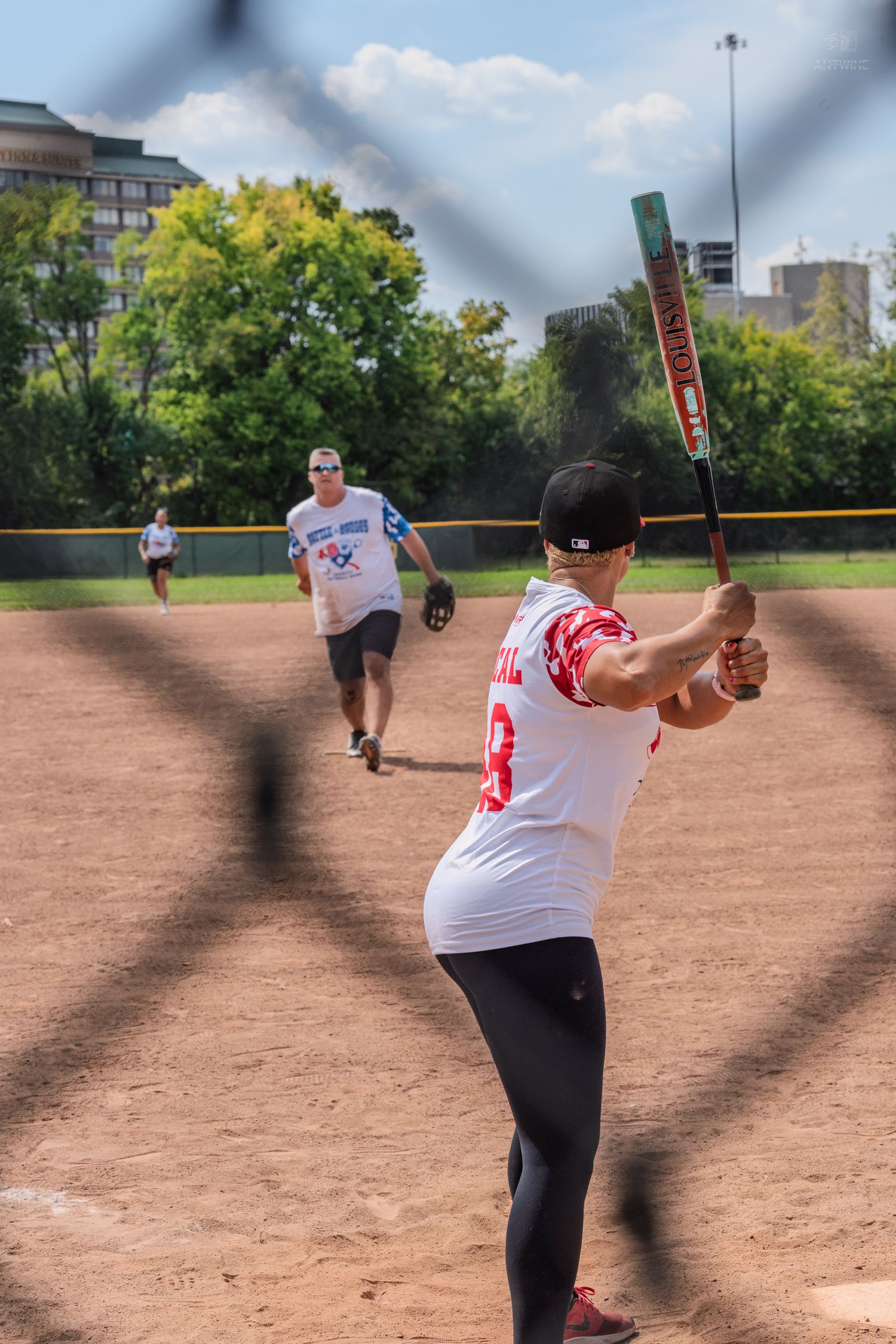 Female softball batter in white uniform preparing to swing, photographed through a chain-link fence.