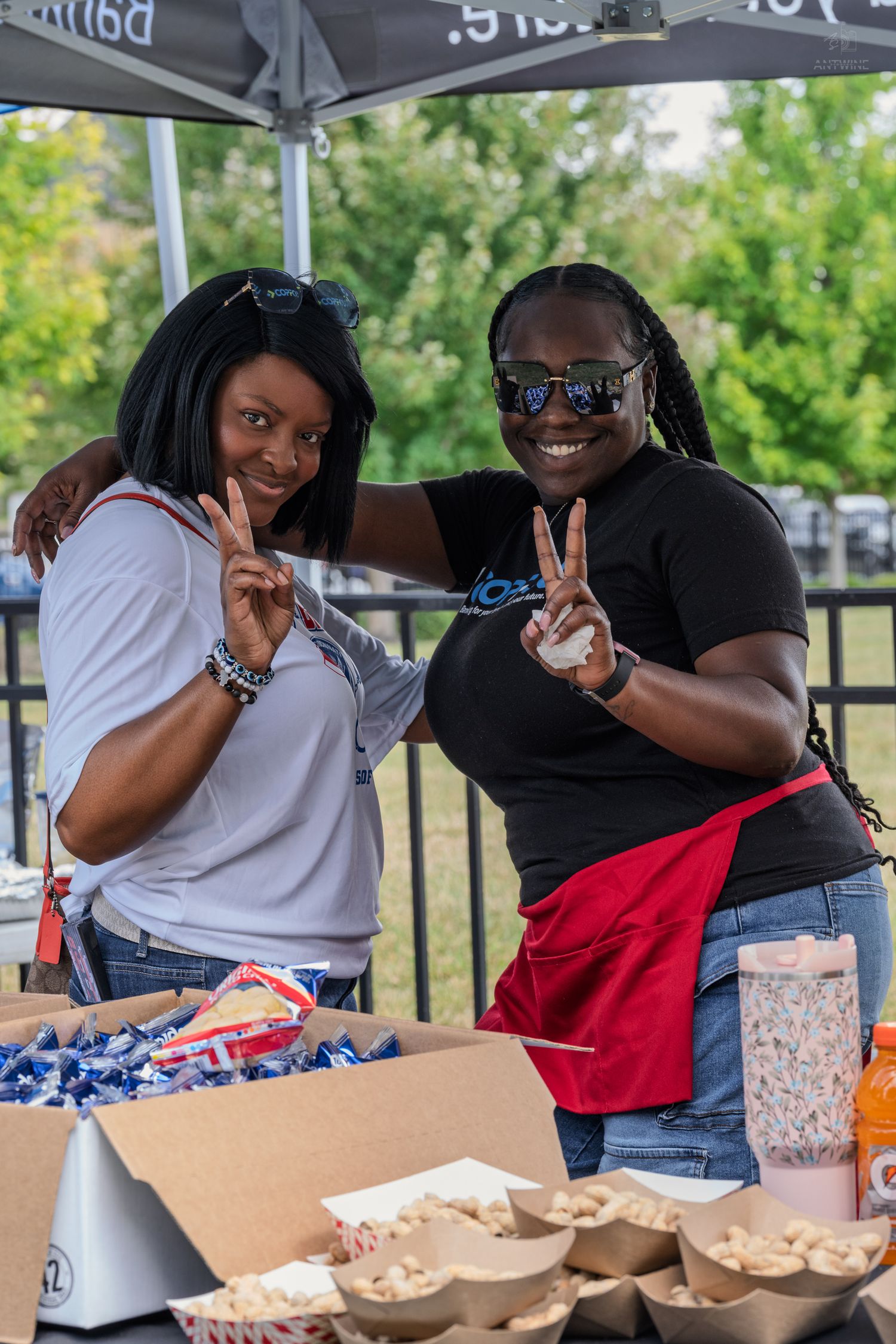 Two women smiling and making peace signs at outdoor charity event food and merchandise table.
