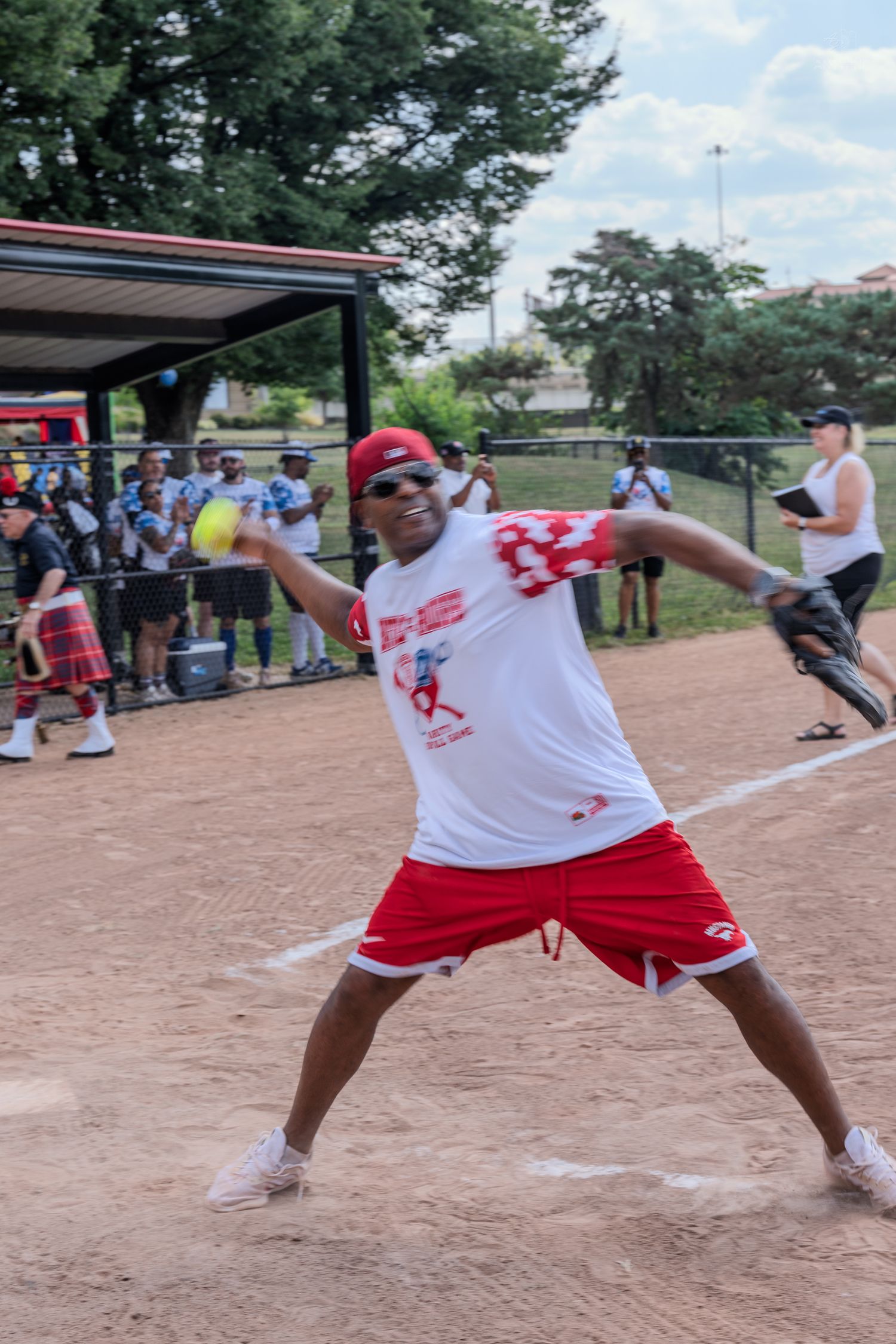 Man in red shorts and white tee pitching a softball on a dirt field during an outdoor charity game.