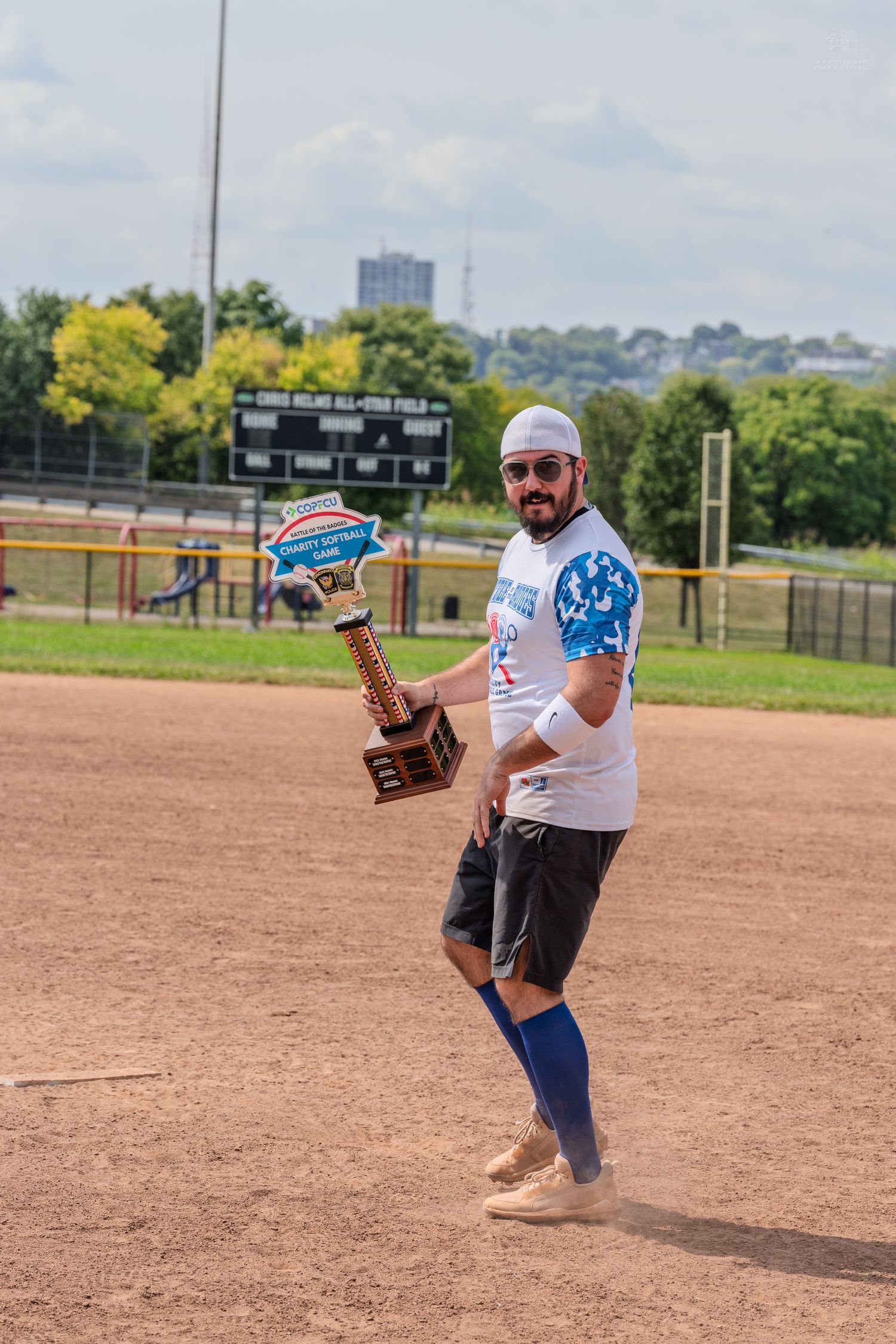 Man in white jersey holds trophy on baseball diamond at Chris Helms All-Star Field on sunny day.